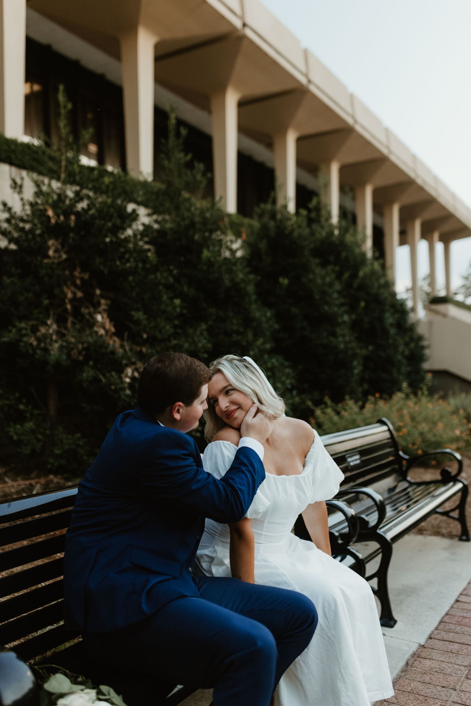 Couple sharing romantic moments on wooden park bench in elegant wedding attire.