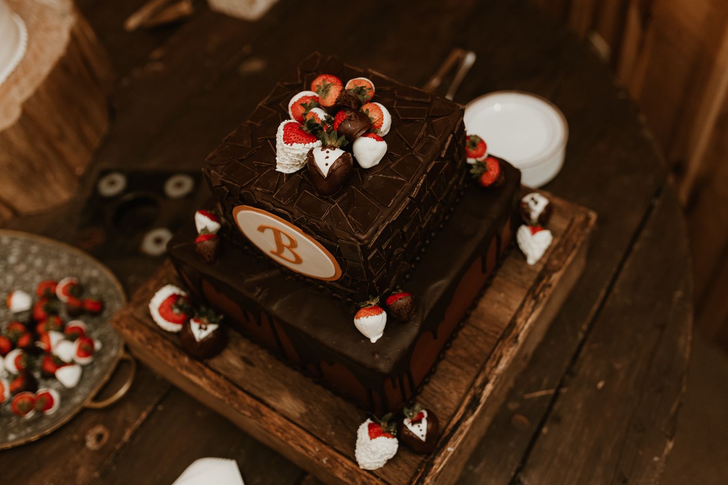 A rustic chocolate wedding cake decorated with chocolate-covered strawberries and white hearts on a wooden table.