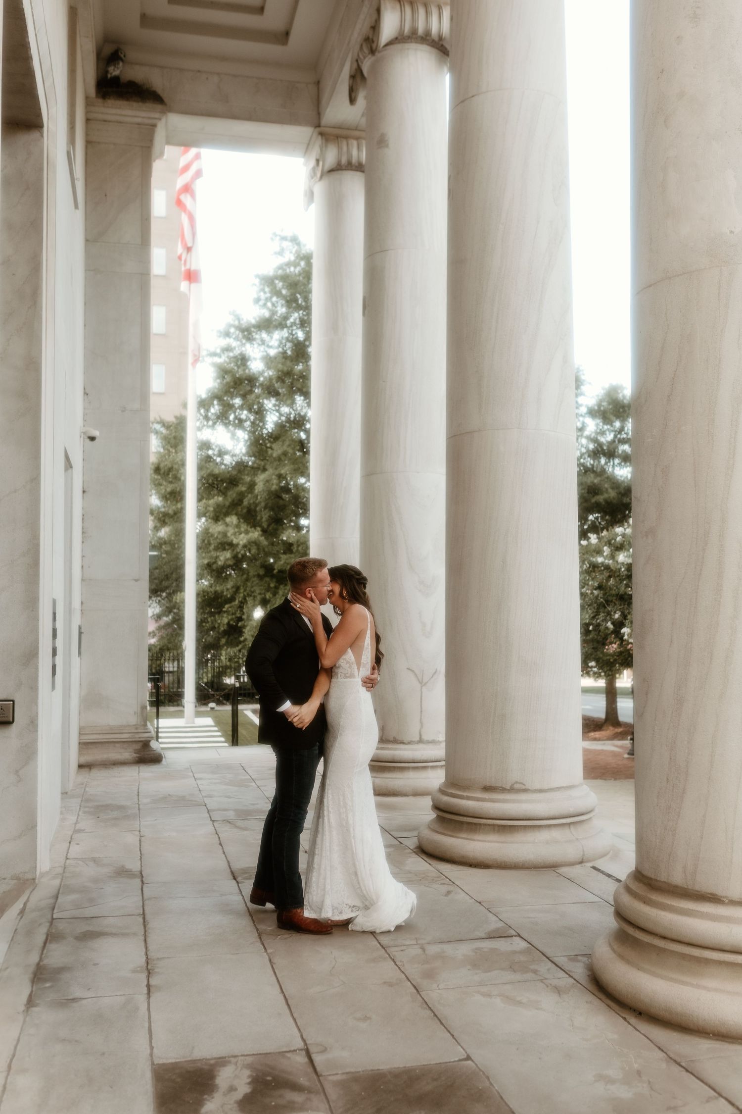 A romantic wedding couple shares an intimate moment between elegant white columns on a grand portico.