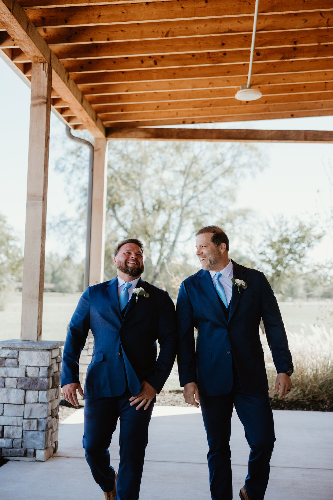 Two grooms in matching navy suits share a joyful moment on a covered porch.