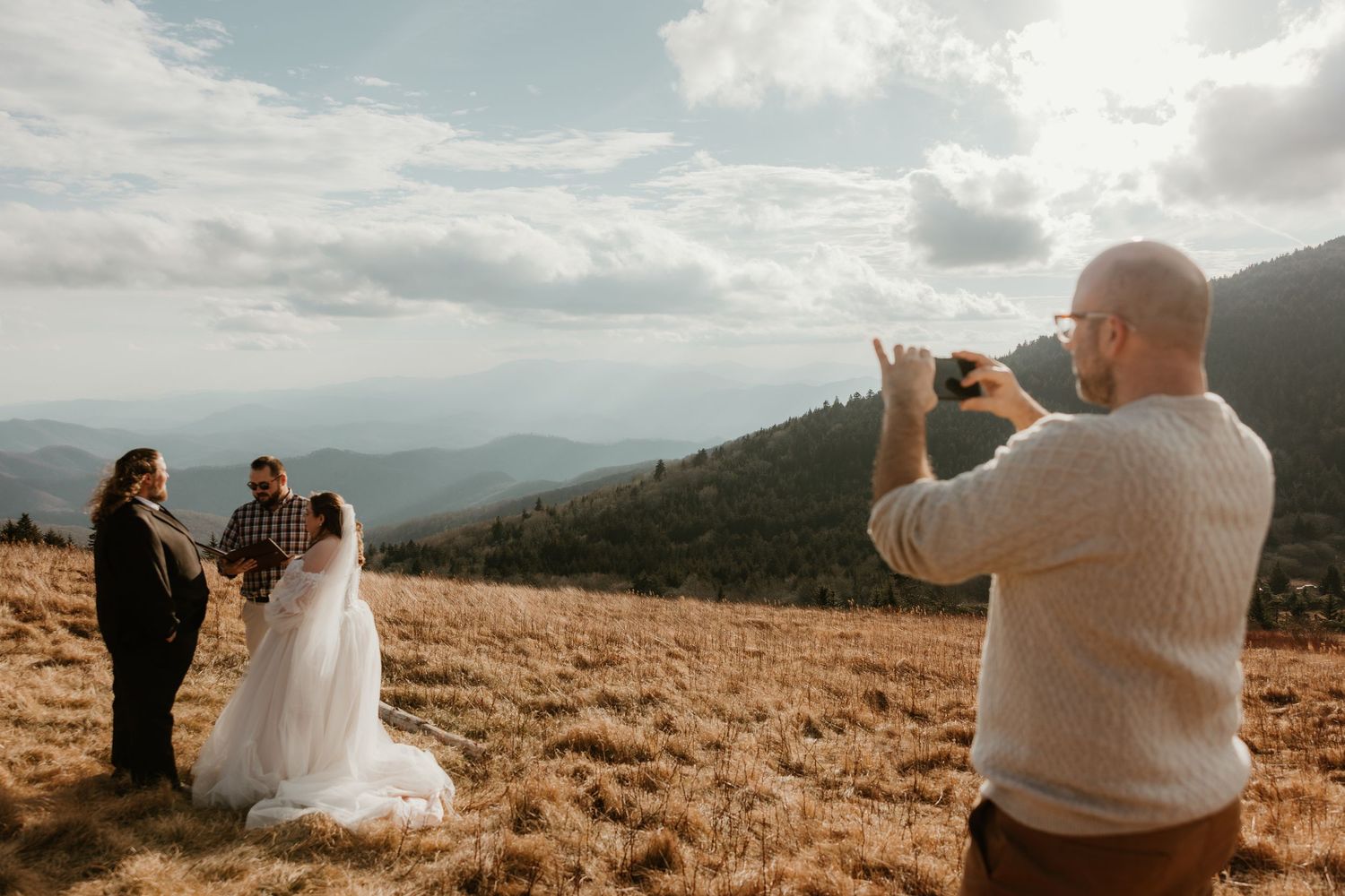 Photographer capturing wedding couple with officiant on scenic mountain meadow under cloudy sky.