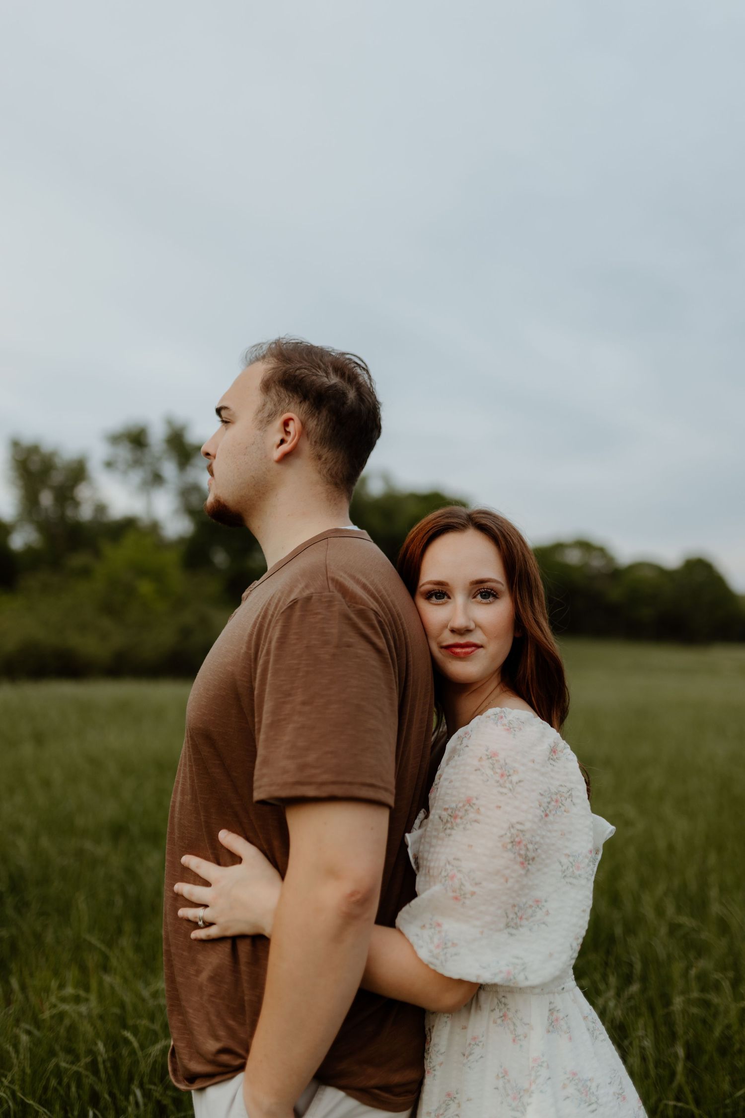 A woman in a white dress embraces a man in a brown t-shirt from behind in a lush green field.