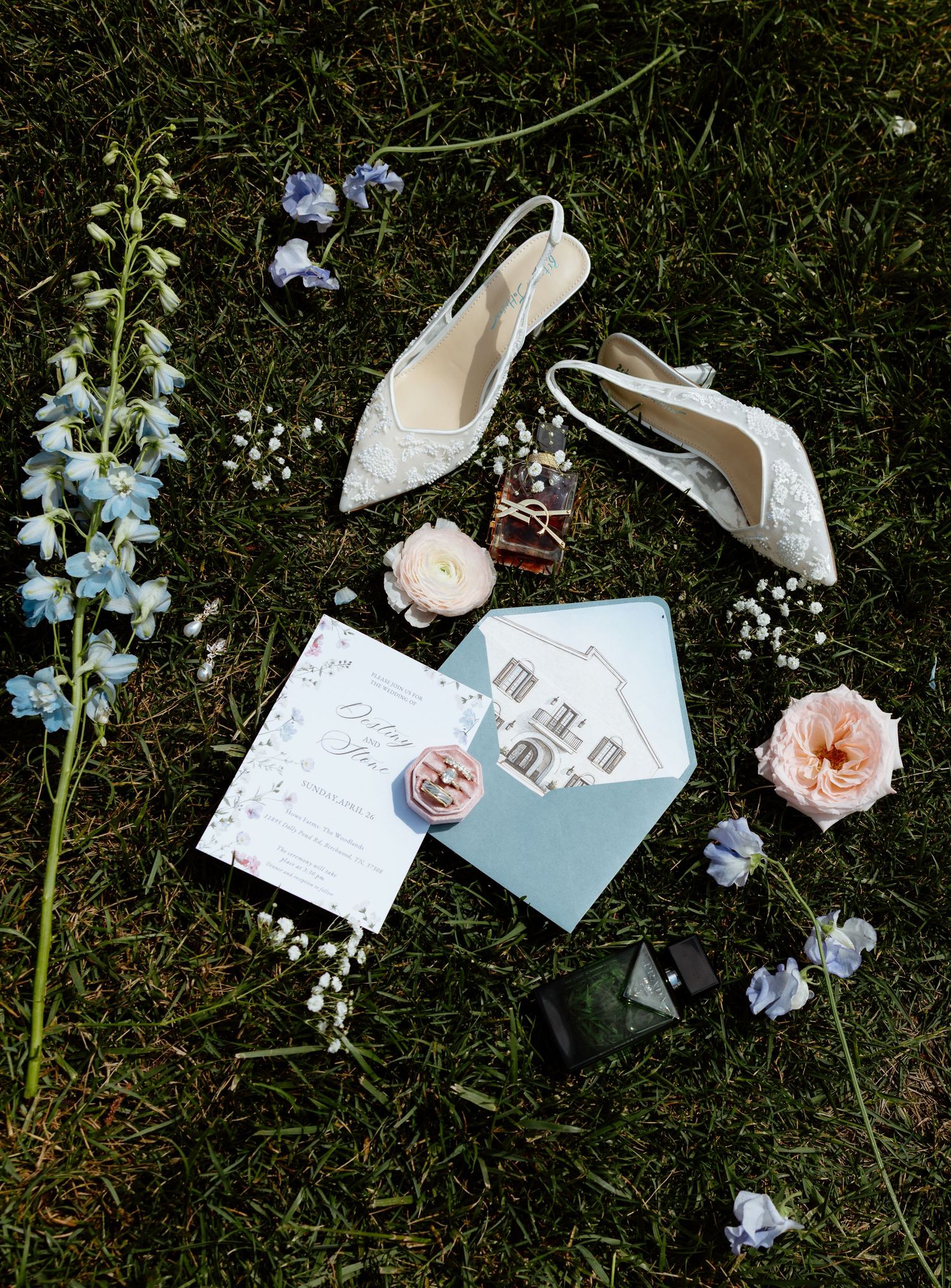 Elegant wedding flat lay on grass featuring lace heels, blue envelope, floral invitations, pink roses, and delicate wildflowers.