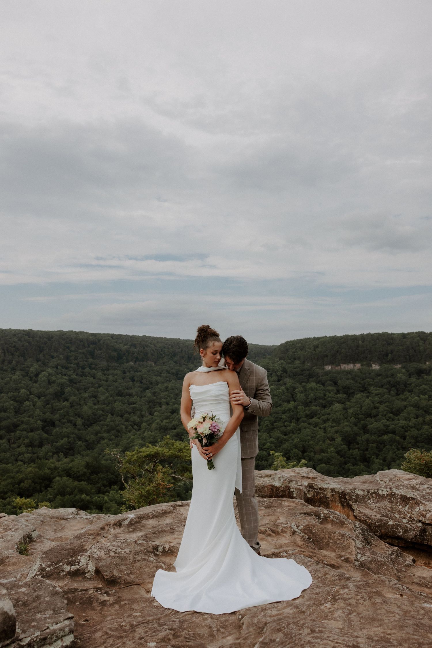 Bride and groom embrace on rocky overlook with lush forest valley backdrop on their wedding day.