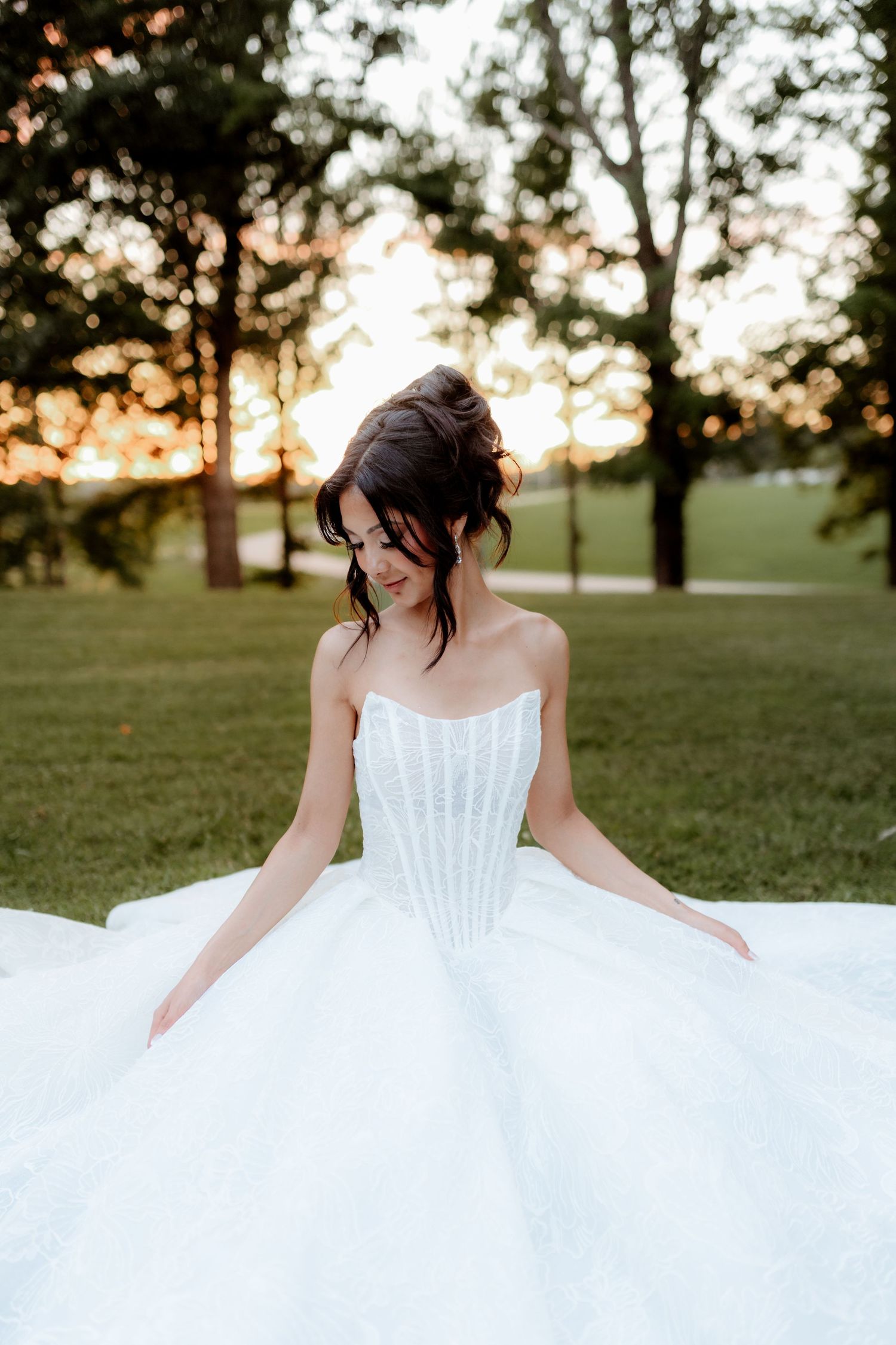Bride in strapless ballgown wedding dress sits outdoors at golden hour sunset surrounded by lush greenery.