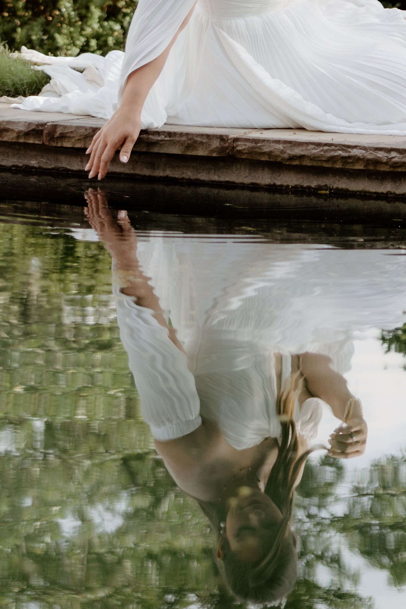 Bride in white gown sitting beside a reflective pond, her elegant dress mirrored in the still water.