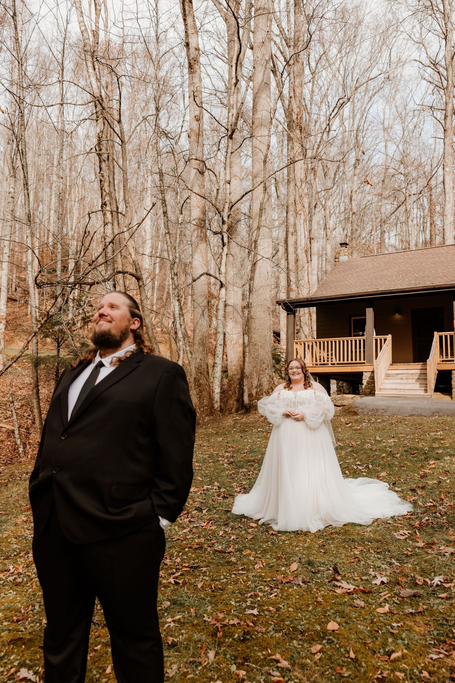 Groom in black suit stands facing away as bride in white gown approaches during first look at woodland cabin wedding.