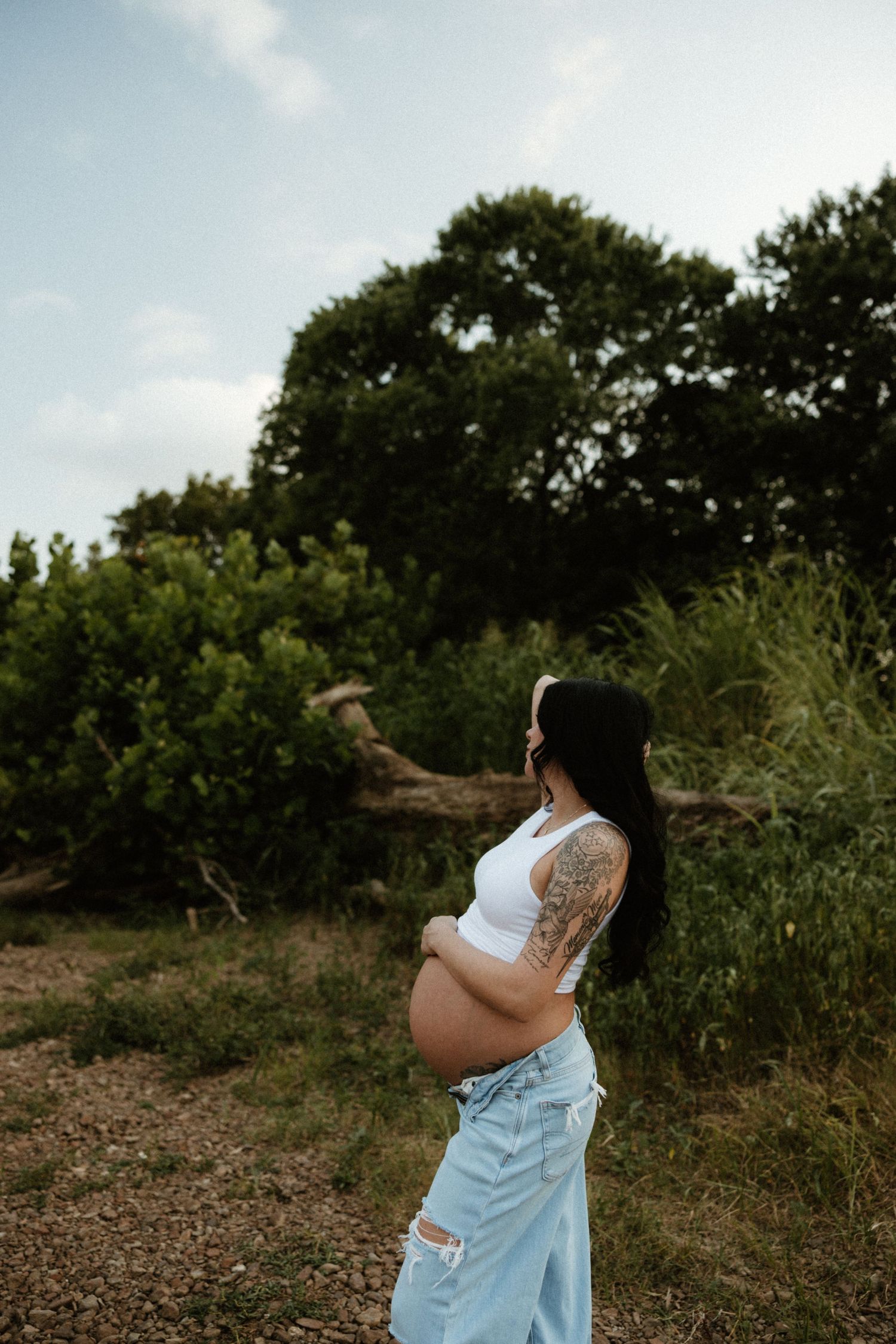 Pregnant person in white tank top and jeans poses outdoors against greenery on a sunny day.