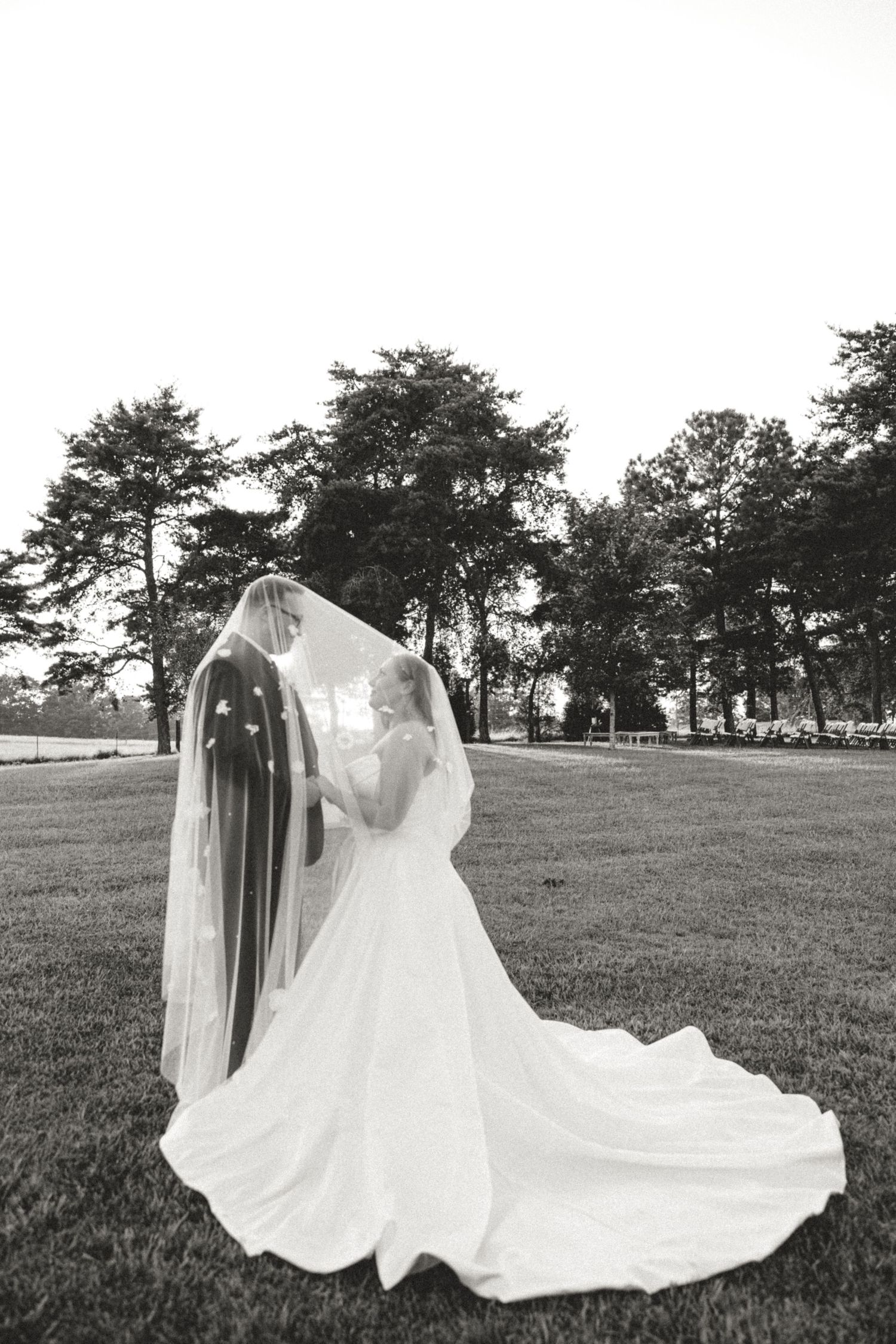 A romantic black and white photograph of a bride in a flowing white wedding gown and veil standing on a grassy lawn.