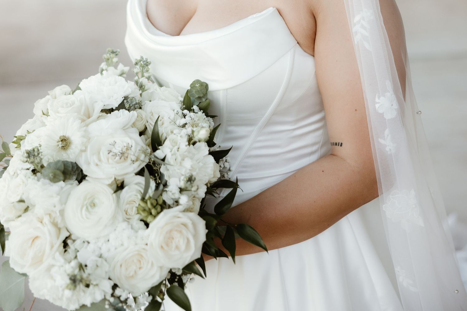 A white wedding bouquet with roses and greenery held by someone wearing a sleeveless white bridal gown and veil.