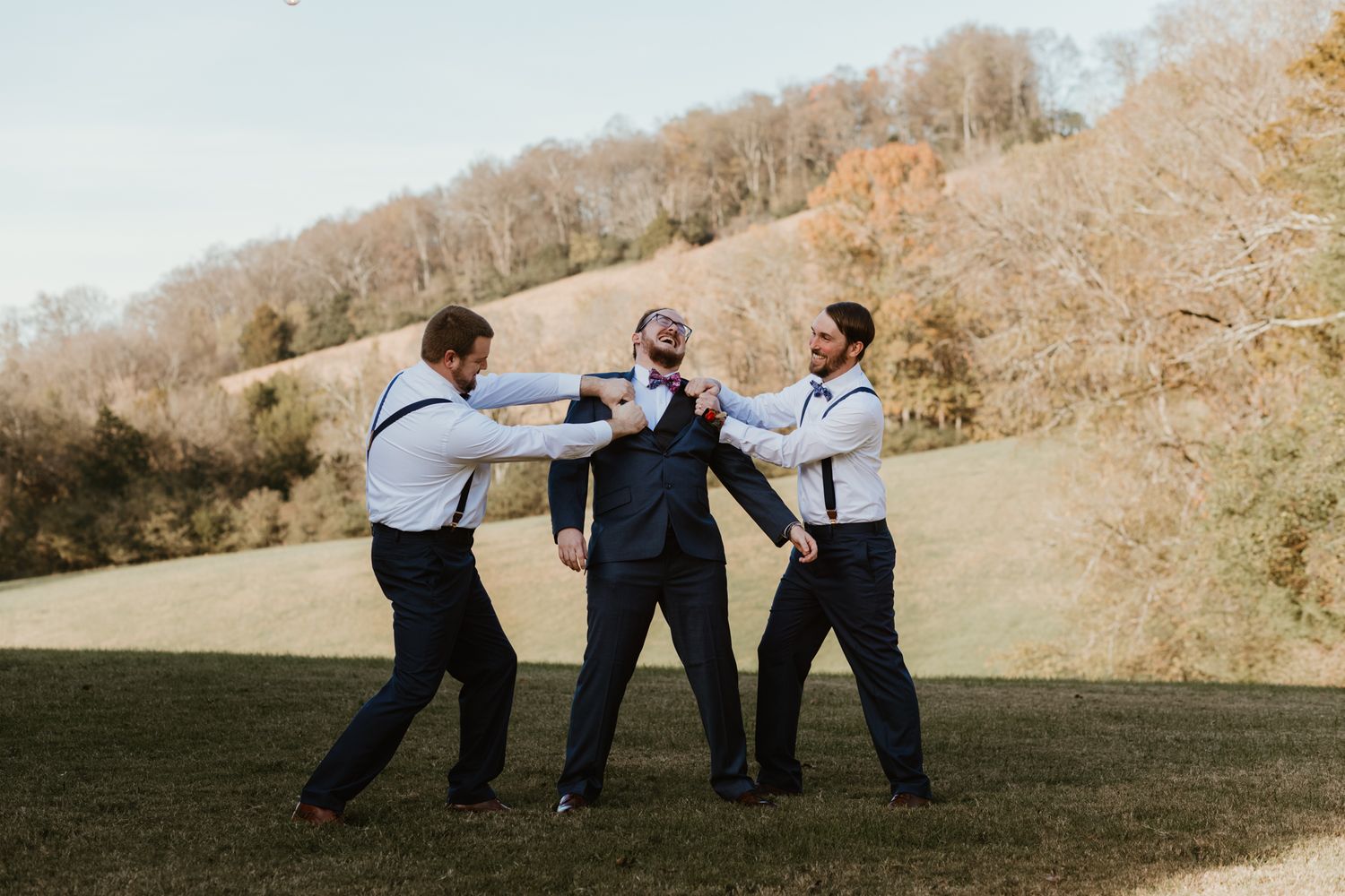 Three groomsmen playfully pose together outdoors on a grassy hillside during a wedding photo session.