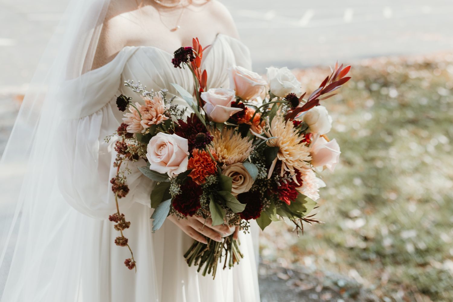 A romantic autumn wedding bouquet with blush roses, burgundy dahlias, and orange accents against an outdoor setting.