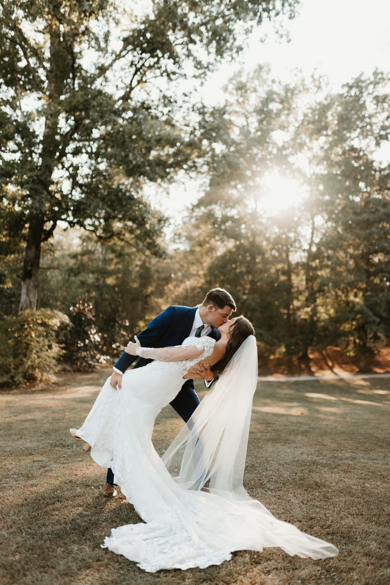 A romantic wedding couple dips in a sunlit forest setting, the bride's long white dress and veil flowing gracefully.