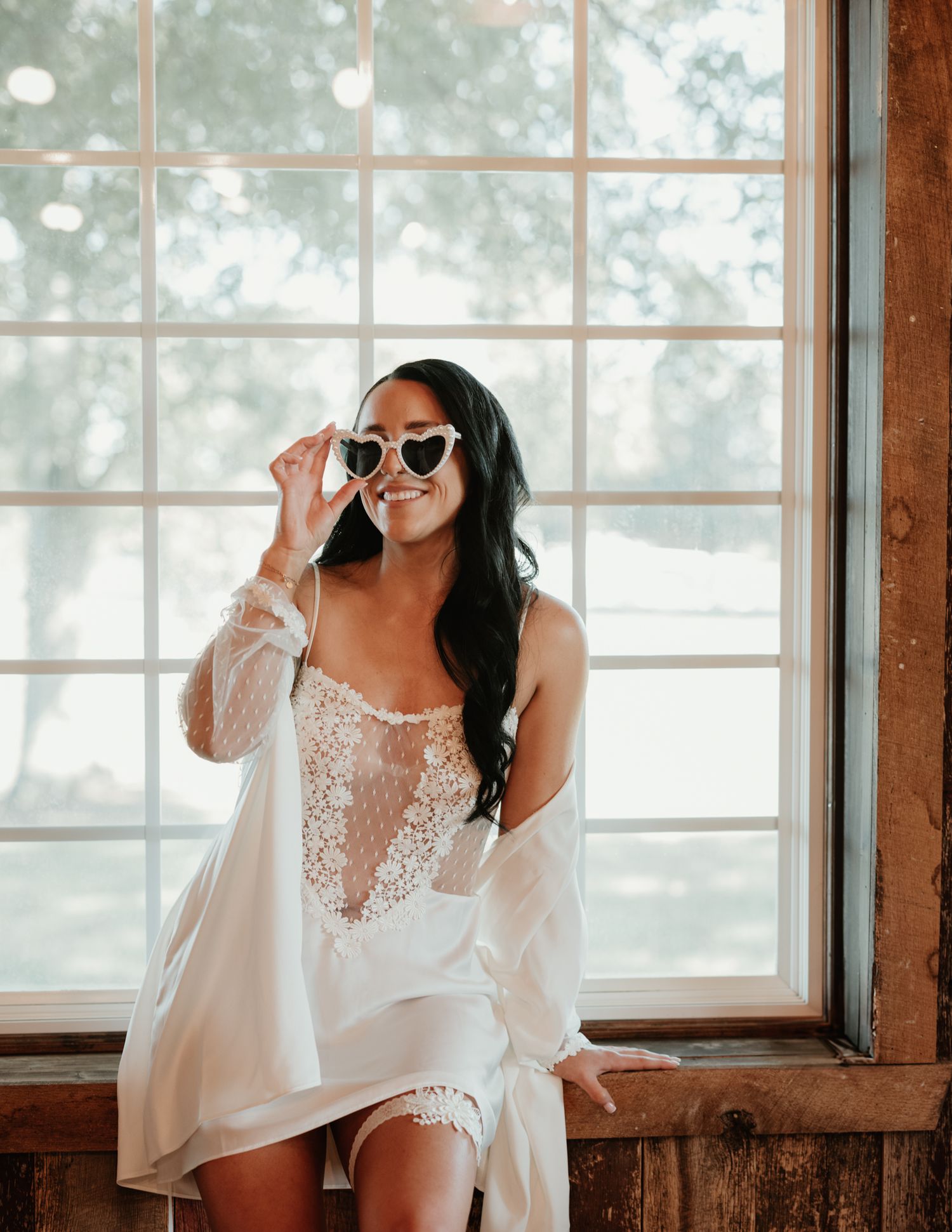 A bride wearing sunglasses and a white robe poses playfully by a large window in natural light.