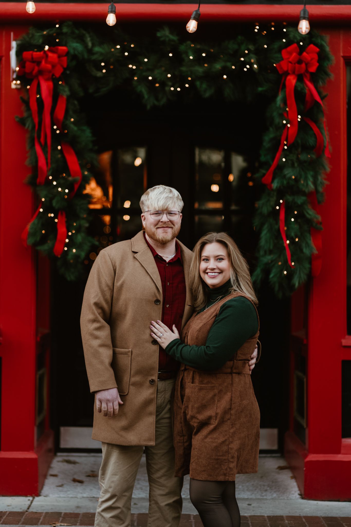 A couple stands together under a festive red archway decorated with holiday bows and twinkling lights.