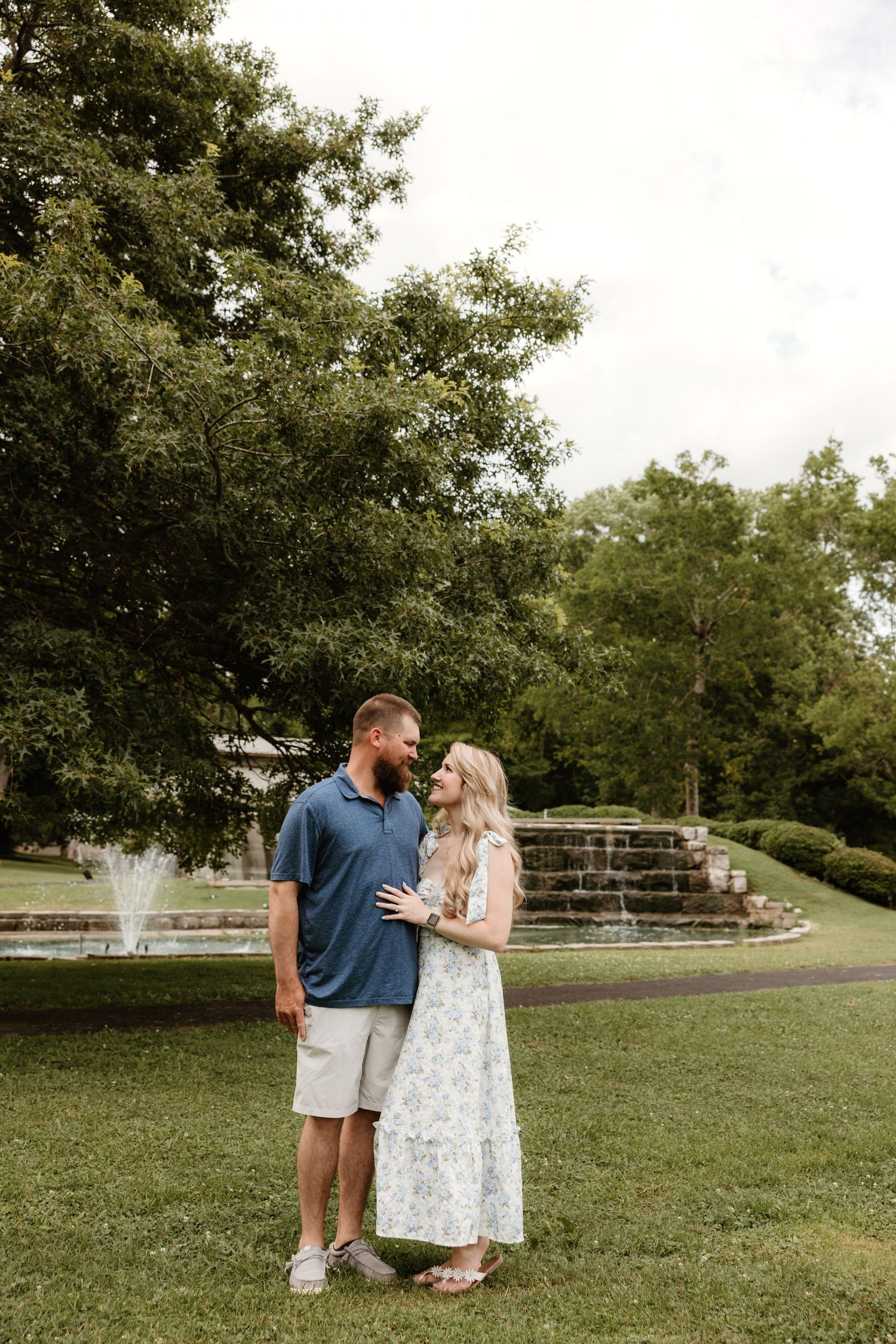 Couple stands together in front of an outdoor water feature on a summer day.