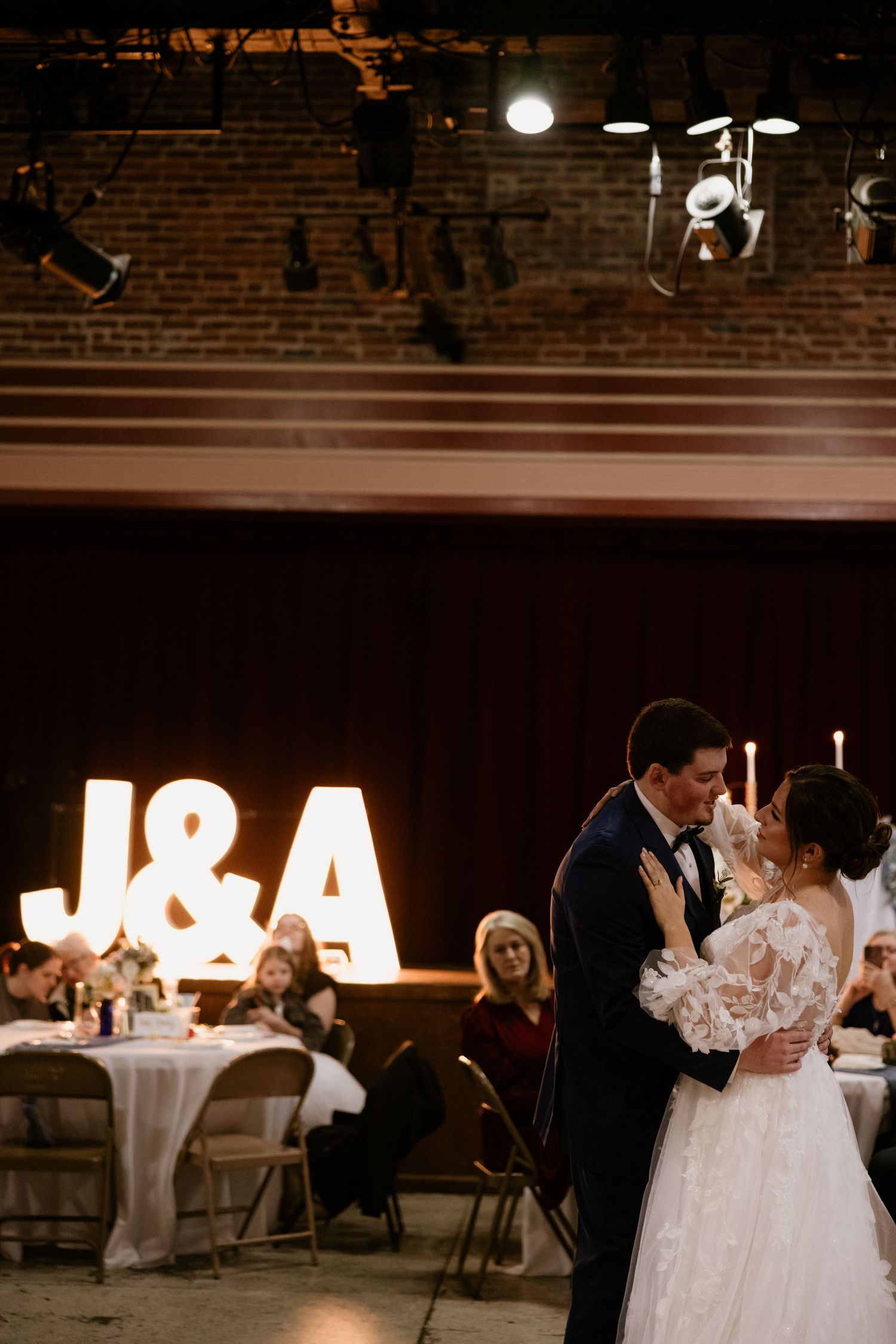 A couple shares their first dance under illuminated J&A letters at a rustic reception venue.