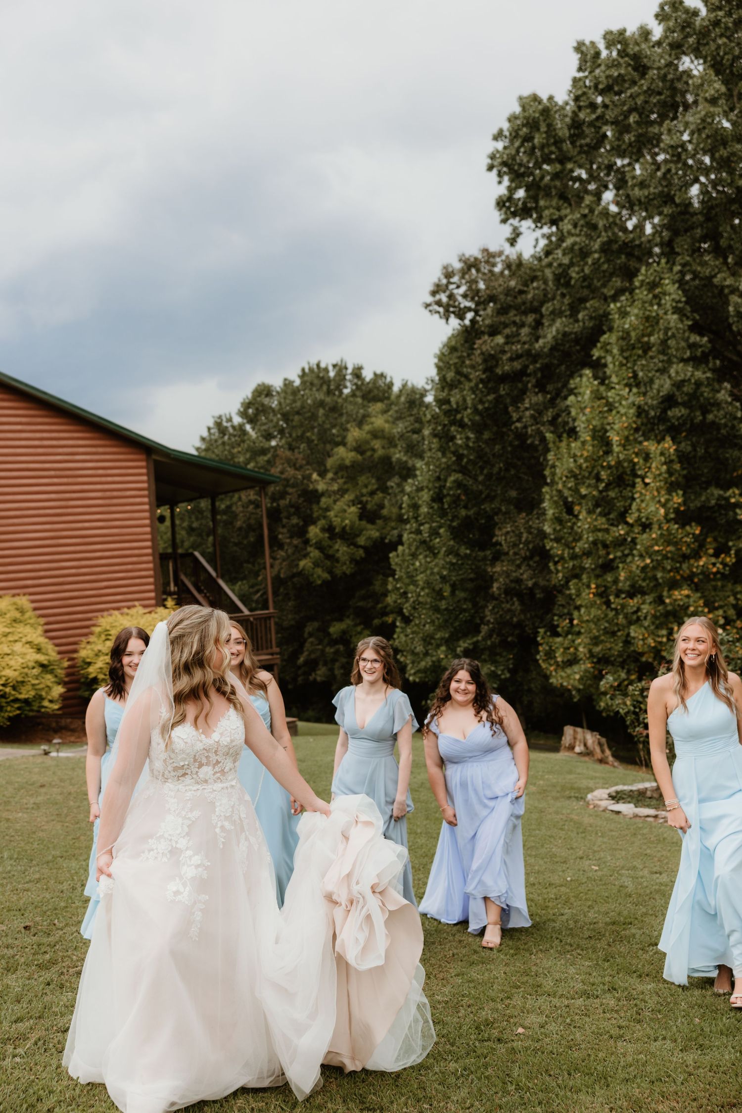 Bridesmaids in light blue dresses walk with the bride in her white gown across a grassy outdoor wedding venue.