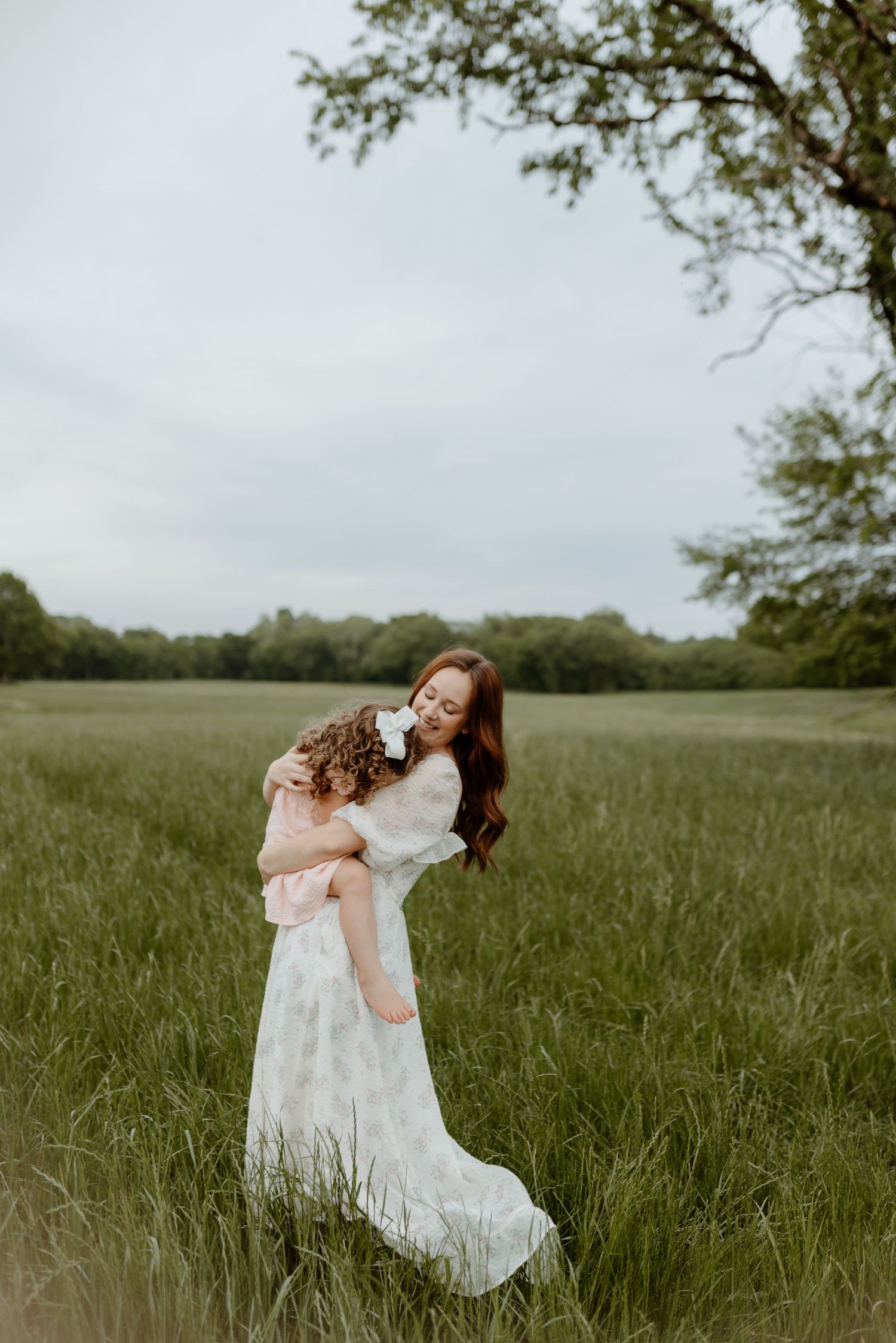 A woman in a white dress holds a young child in a lush green field under a cloudy sky.