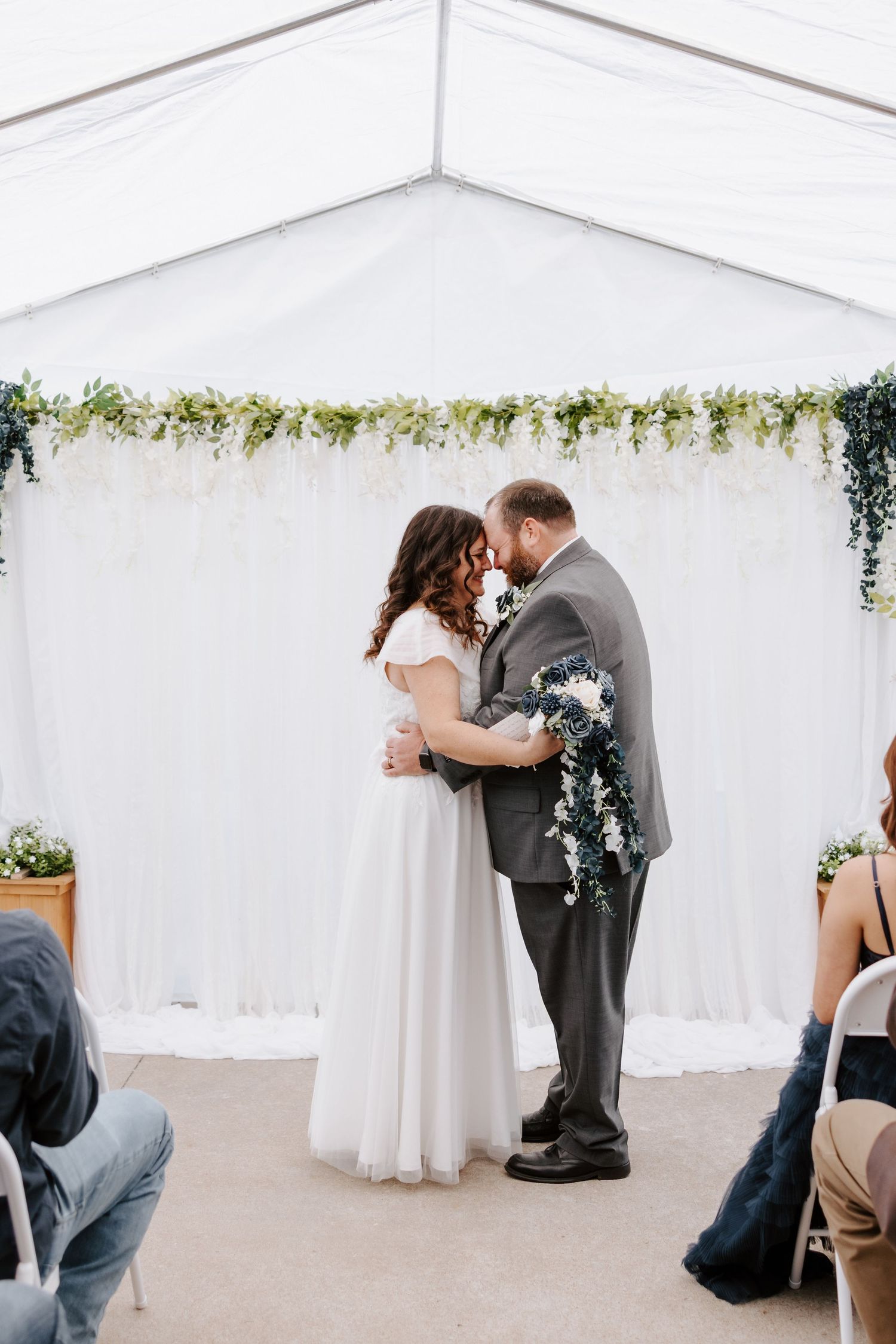 A bride and groom share their first kiss at a romantic outdoor wedding ceremony under a white tent with floral greenery backdrop.