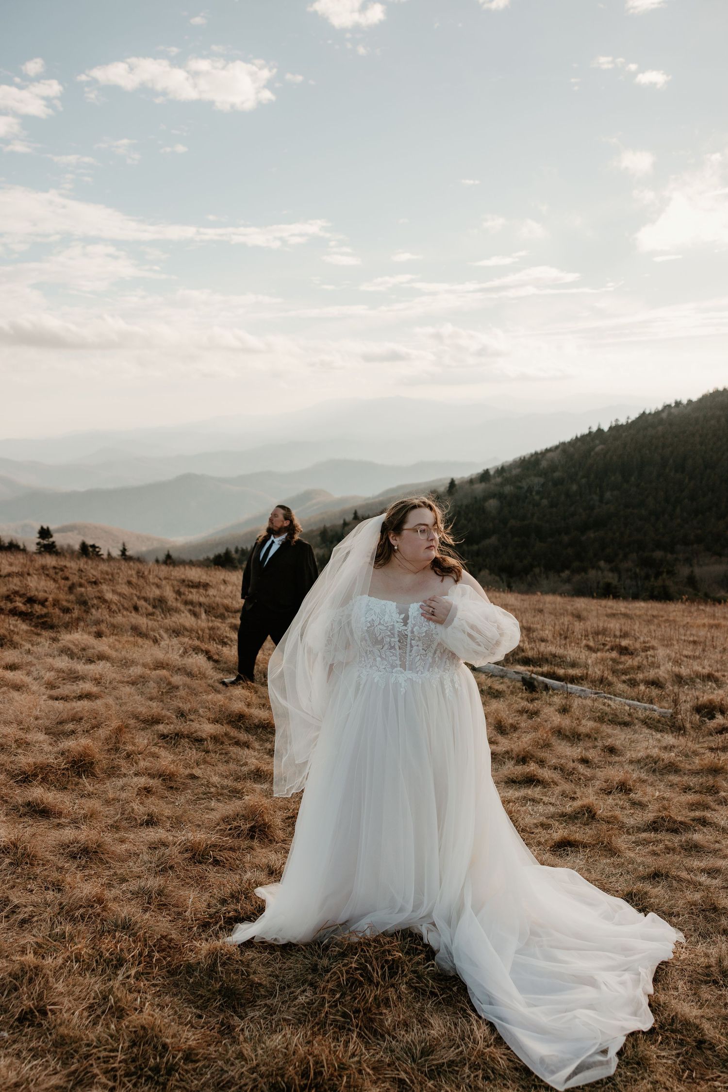 Bride in flowing white gown and veil stands on mountain meadow with groom in black suit and misty peaks behind.
