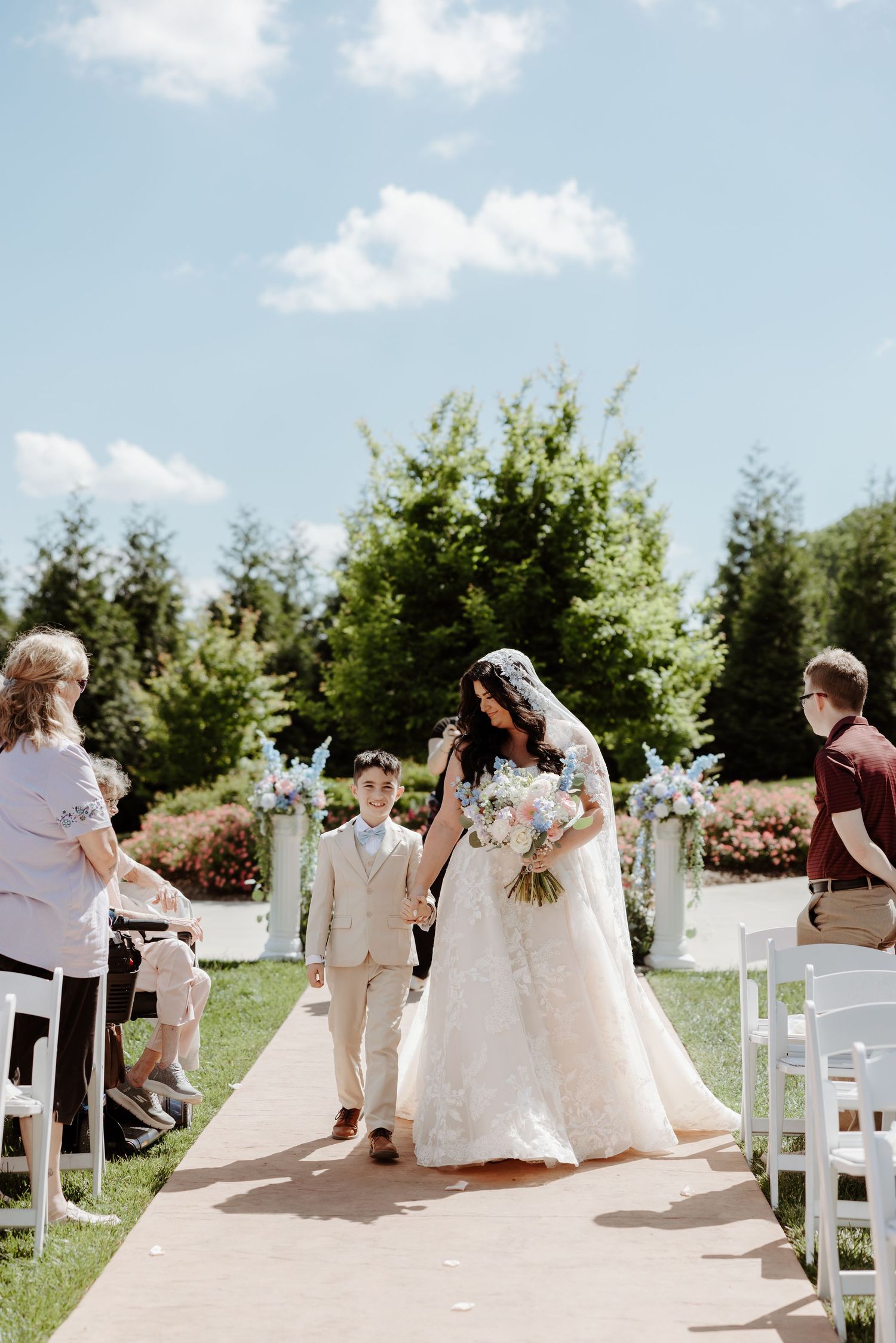 Bride in lace ball gown walks down outdoor aisle with young boy on sunny wedding day.