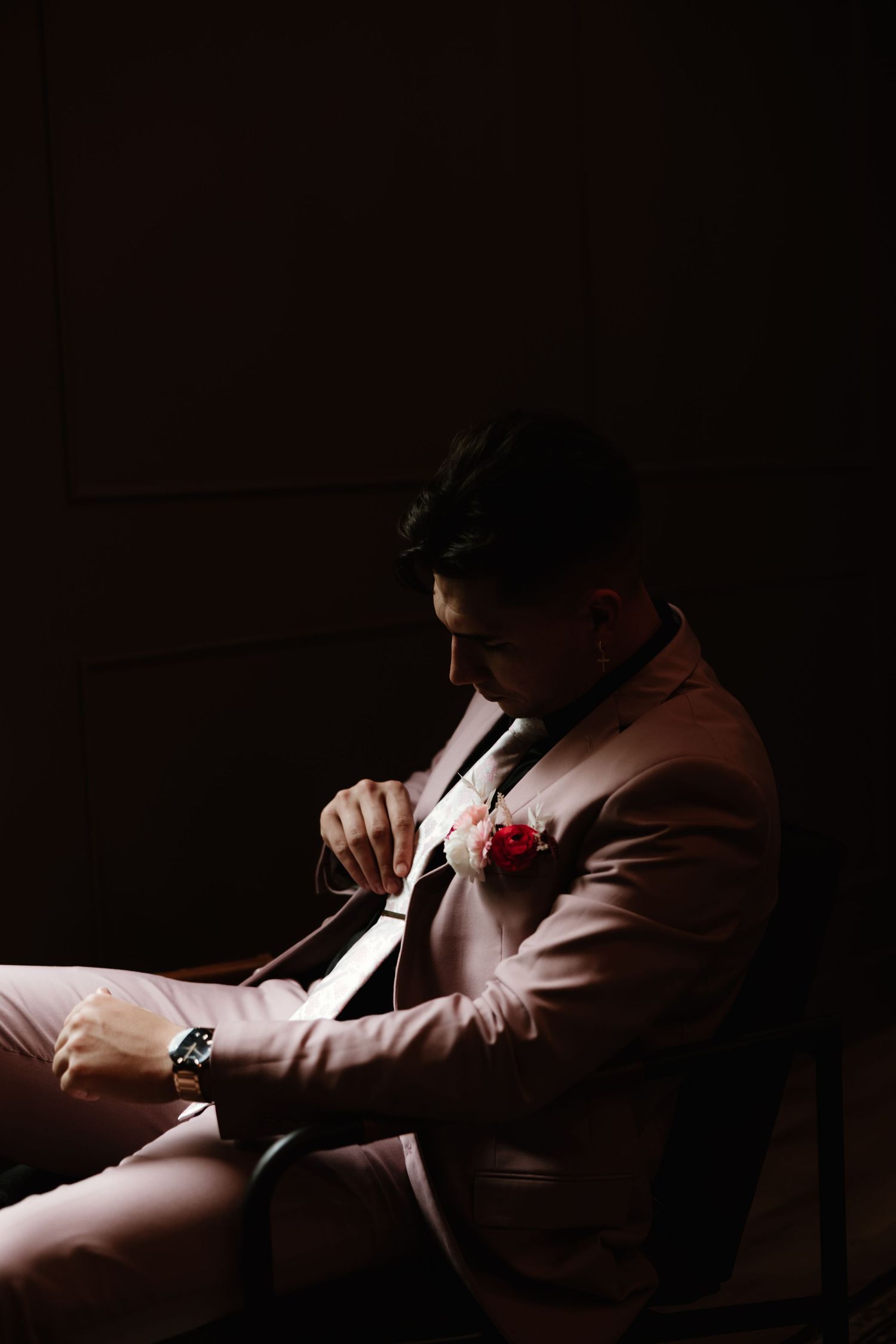 A stylish groom in a brown suit jacket adjusts his tie while seated in dramatic low-key lighting.