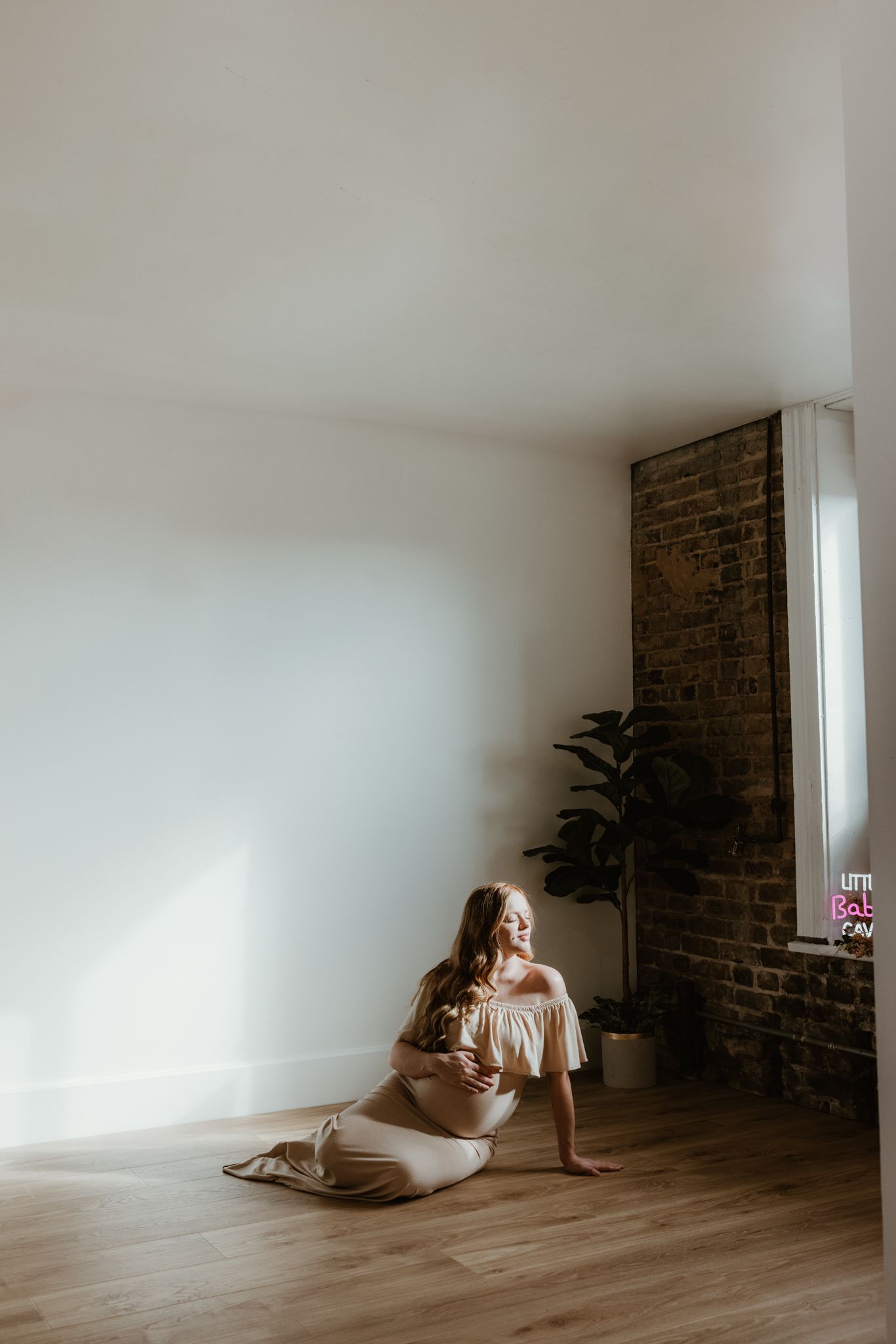 A person sits on wooden floor beside exposed brick wall in minimalist room with natural light.