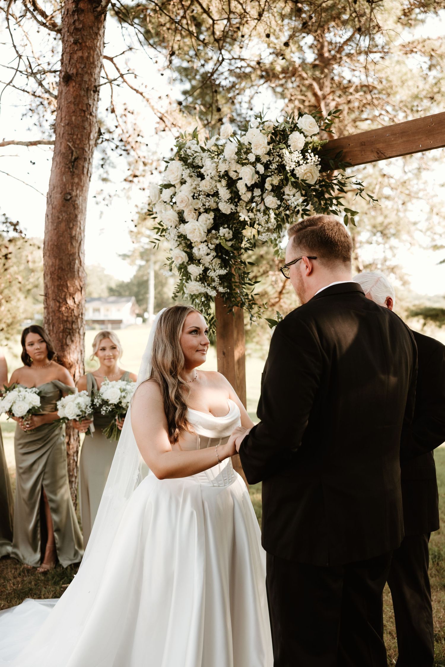A romantic outdoor wedding scene with bride in flowing white dress and groom in black suit under sunlit trees.