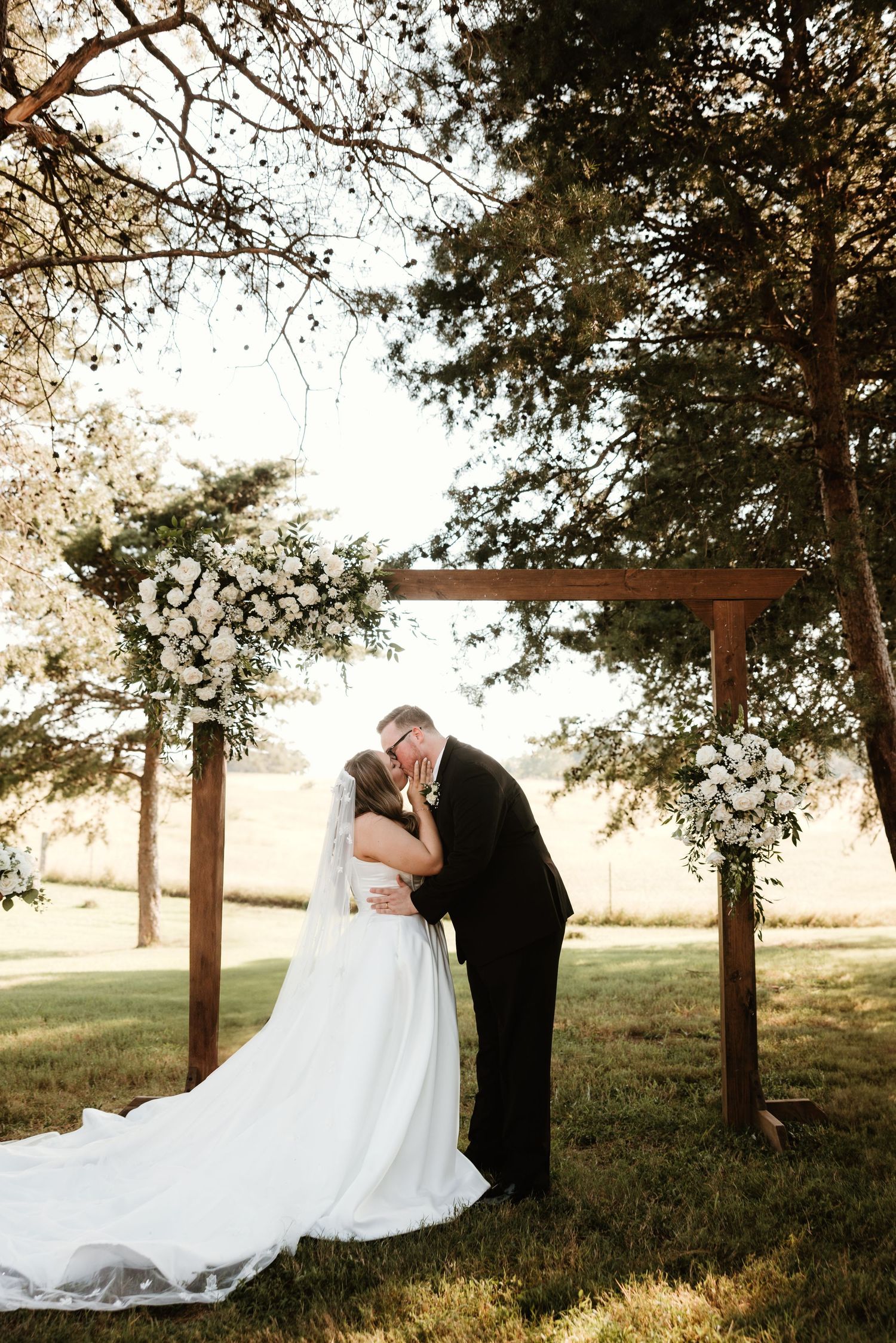 A couple shares an intimate wedding moment under a wooden arch at sunset in a natural outdoor setting.