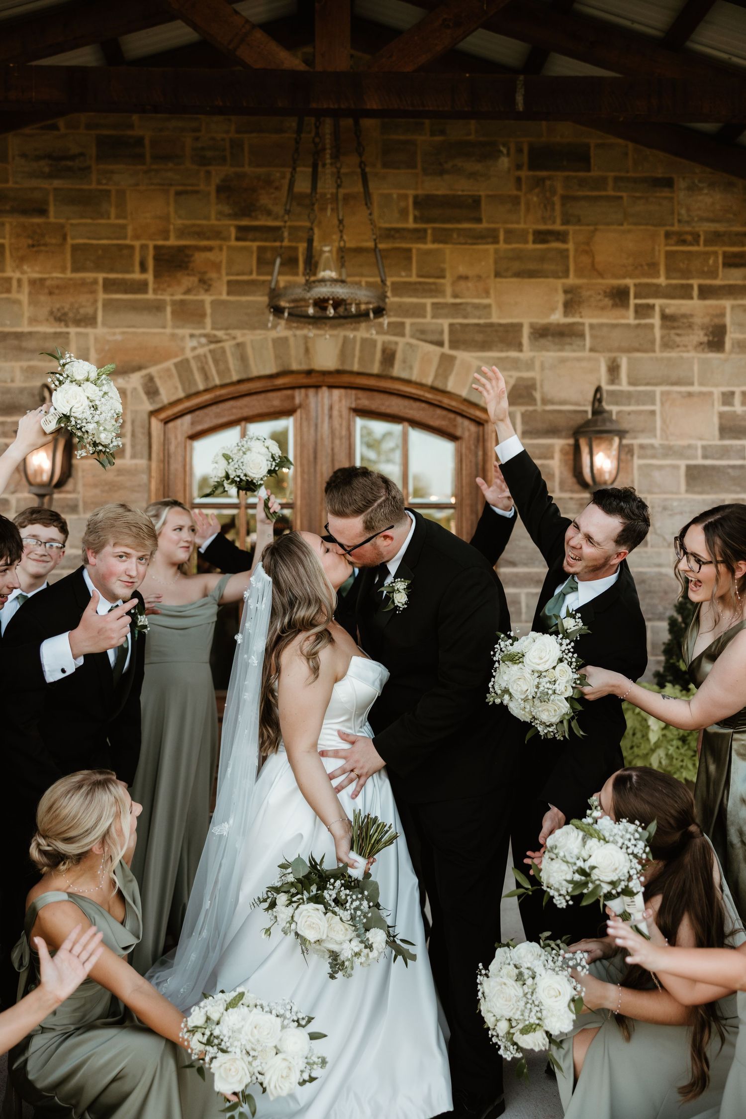 Joyful wedding party celebrates with white floral bouquets against stone wall backdrop during intimate outdoor ceremony.