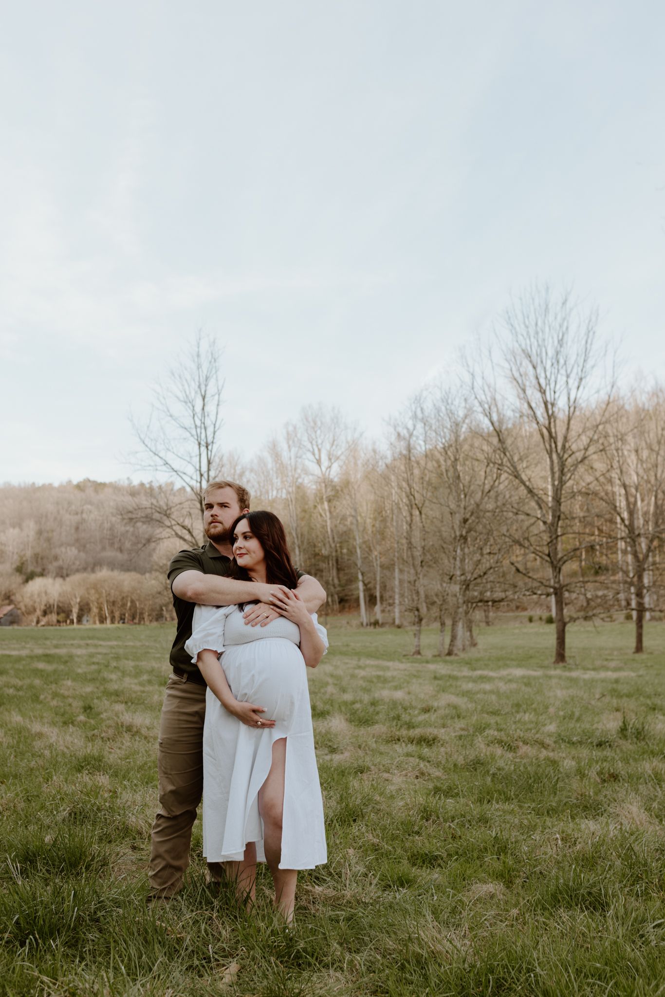 A couple embraces in a natural outdoor setting with bare trees and grass during maternity photoshoot.