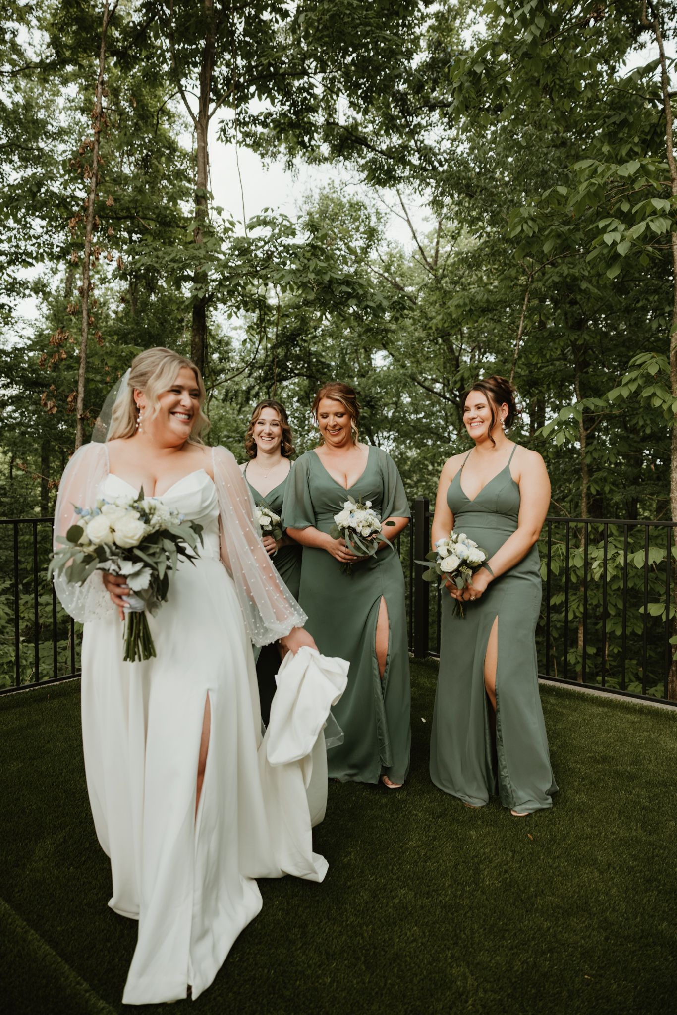 A bridal party in elegant sage green dresses holding white bouquets pose together on a grassy outdoor setting.
