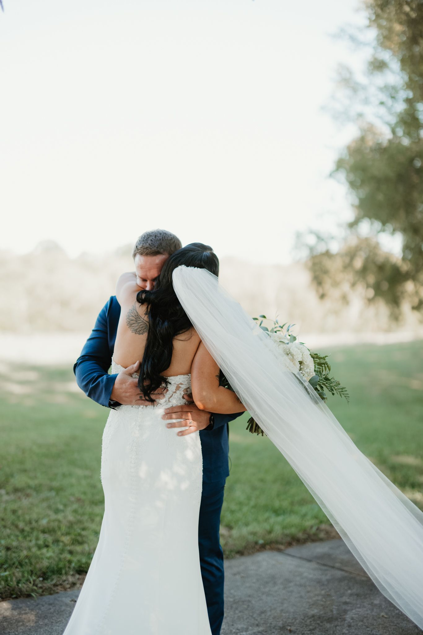 A romantic wedding moment captured as a couple embraces on a sunlit path with a flowing veil in the breeze.
