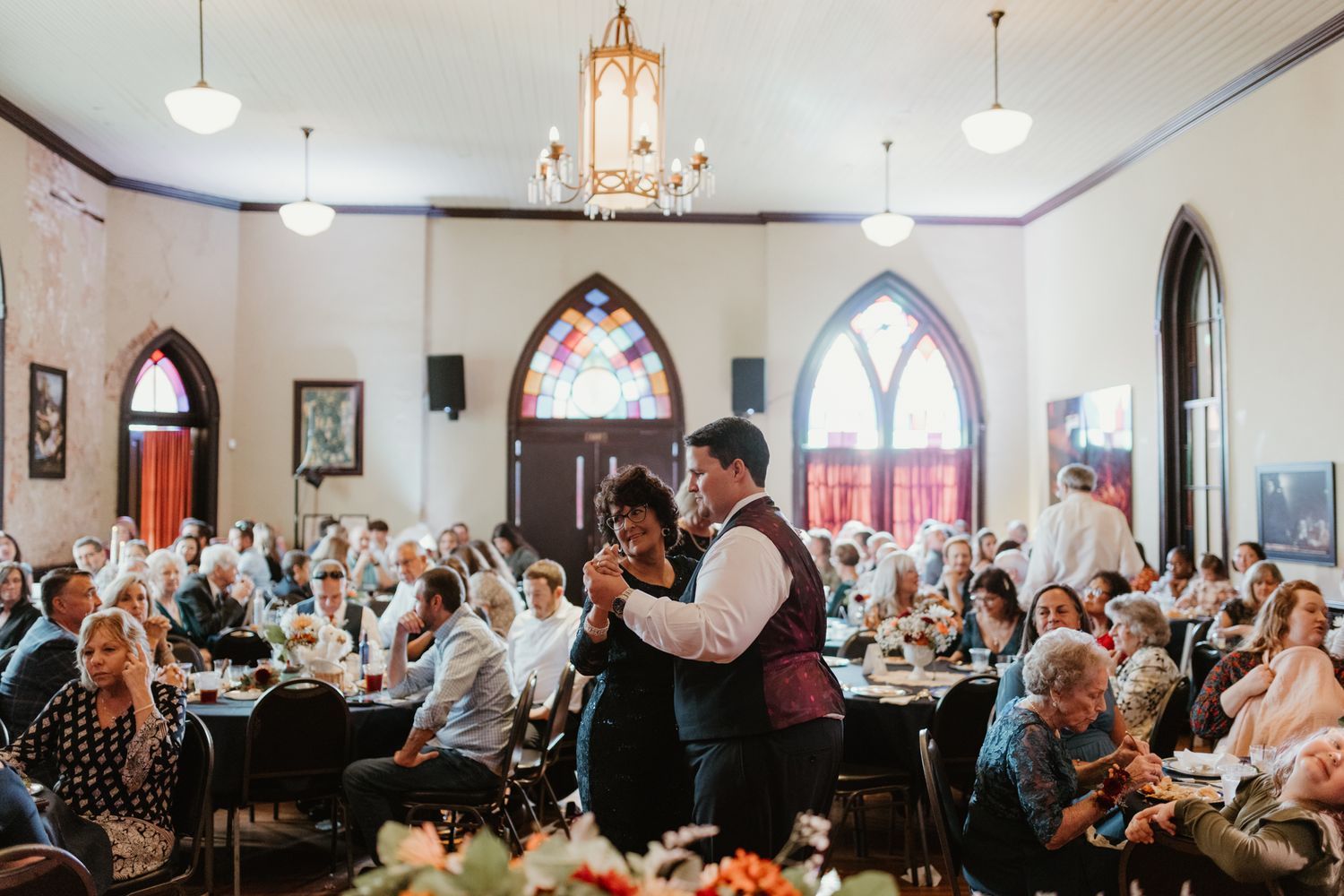Guests gather inside an elegant historic church dining hall with chandeliers and arched windows for a community meal service.
