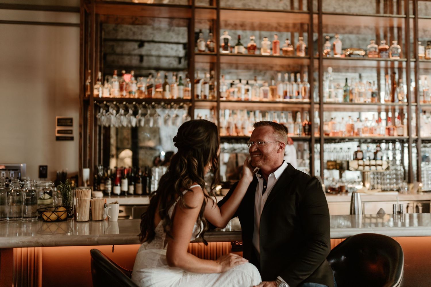 Wedding couple shares intimate moment at modern bar with glowing amber lights and glass shelving in background.