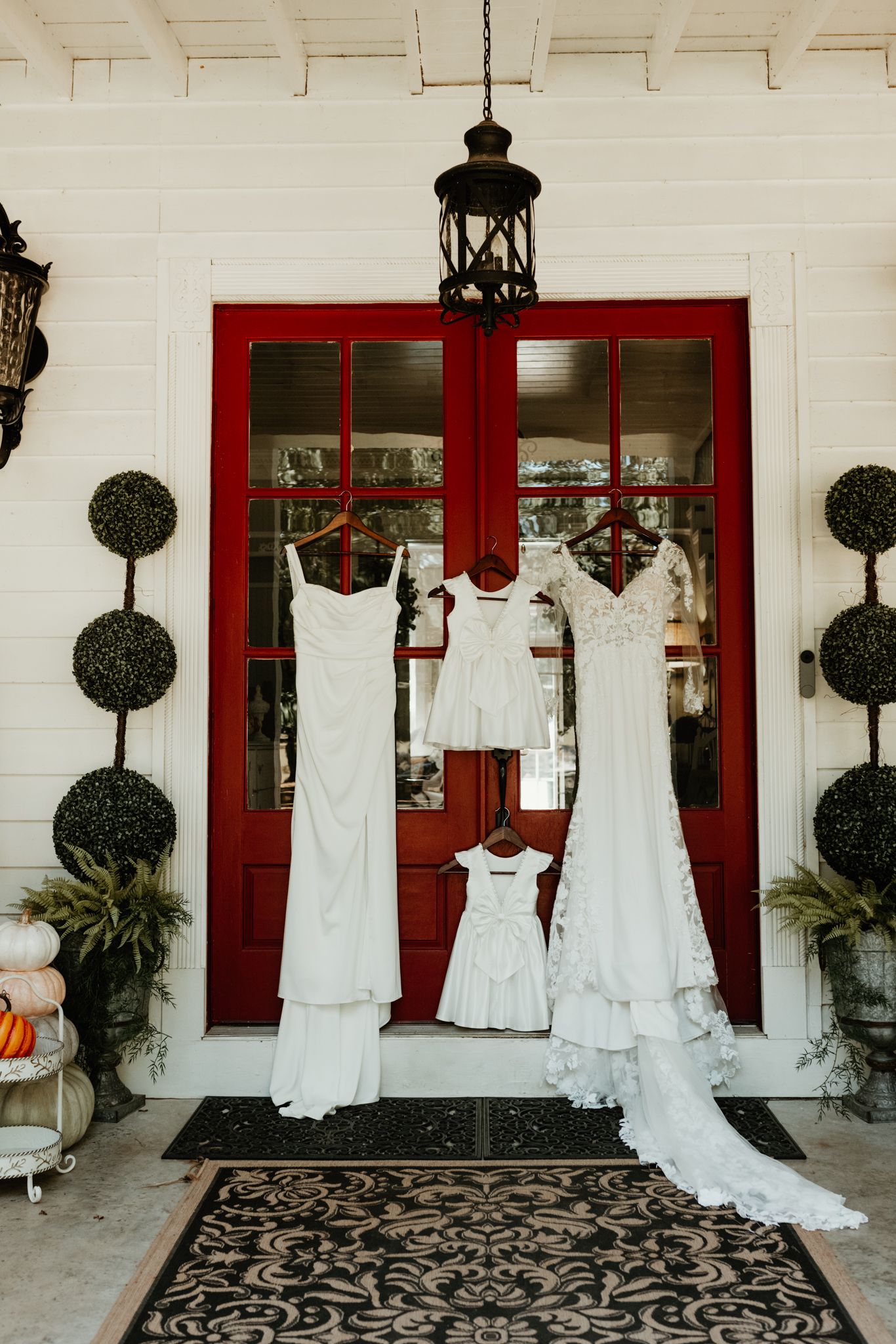 Multiple white wedding dresses and gowns displayed on hangers against red doors of a white building with decorative topiaries.
