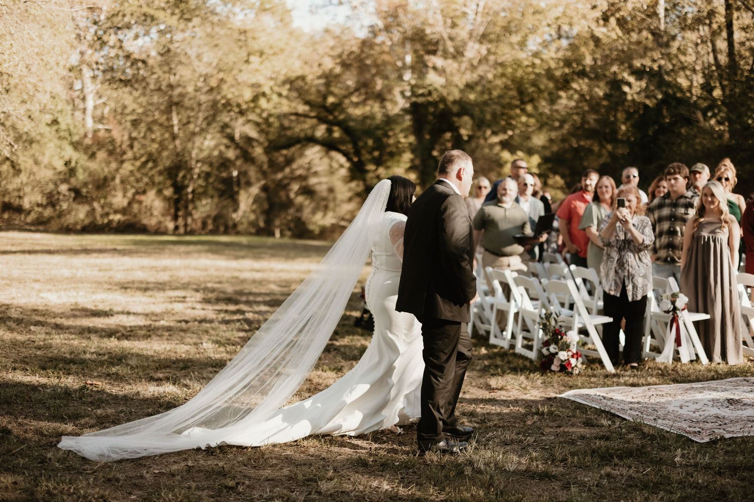 A wedding couple embraces outdoors during their ceremony while guests watch from a sunny wooded setting.