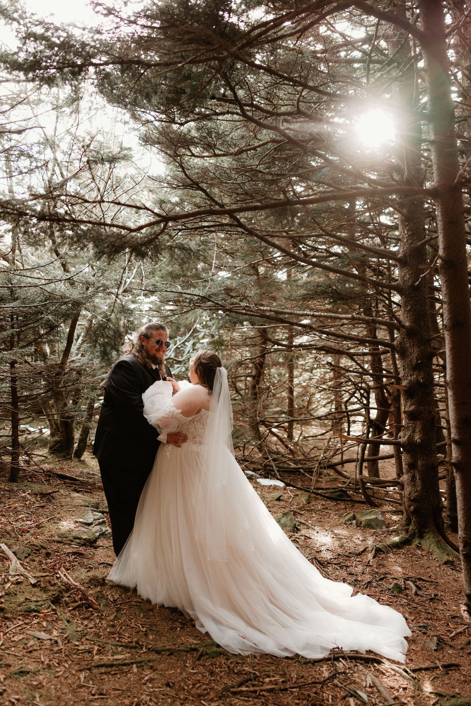 Bride in white gown and groom in black suit embrace in a sunlit forest during their woodland wedding ceremony.