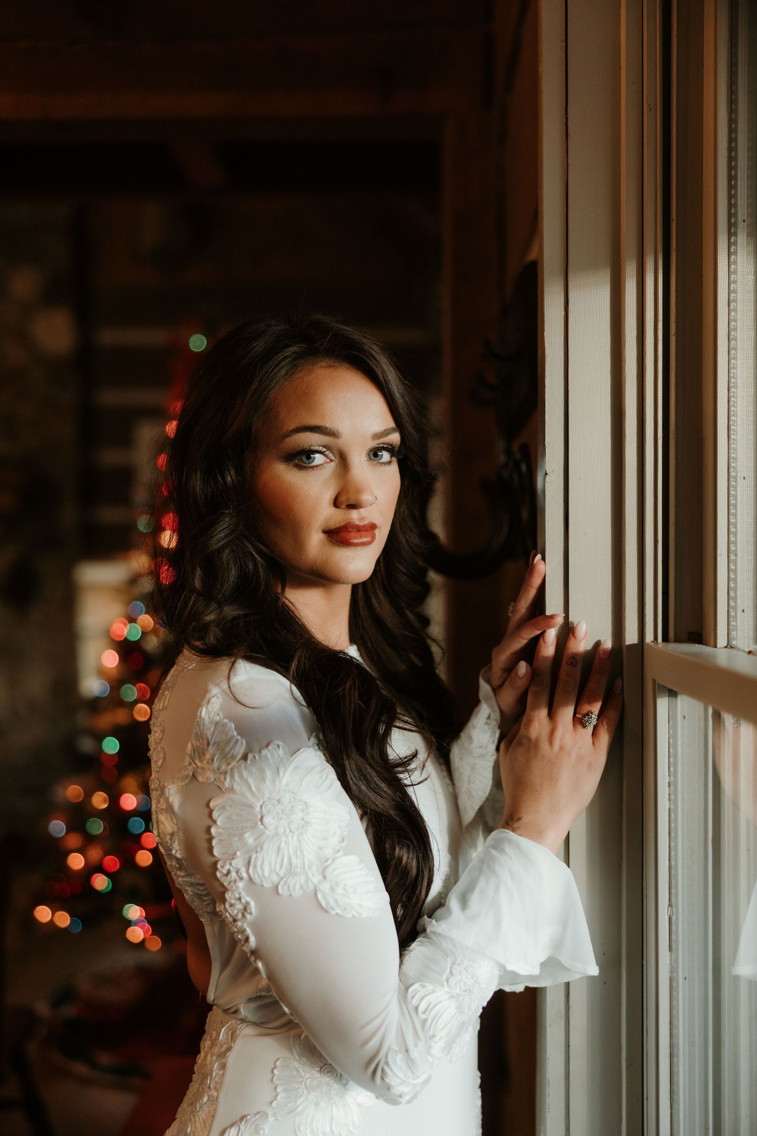 Bride in white lace long-sleeve gown poses by window with Christmas tree bokeh lights in background.