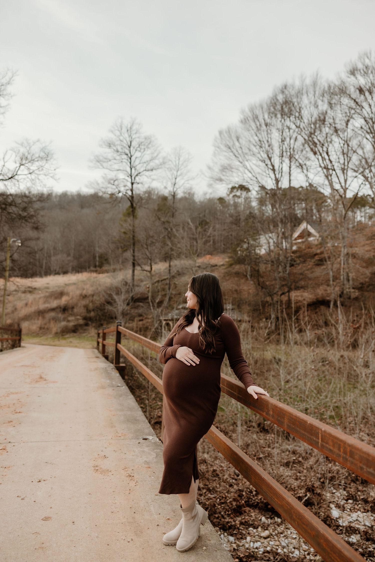Pregnant woman in brown dress on wooden bridge, cradling baby bump, surrounded by winter trees.