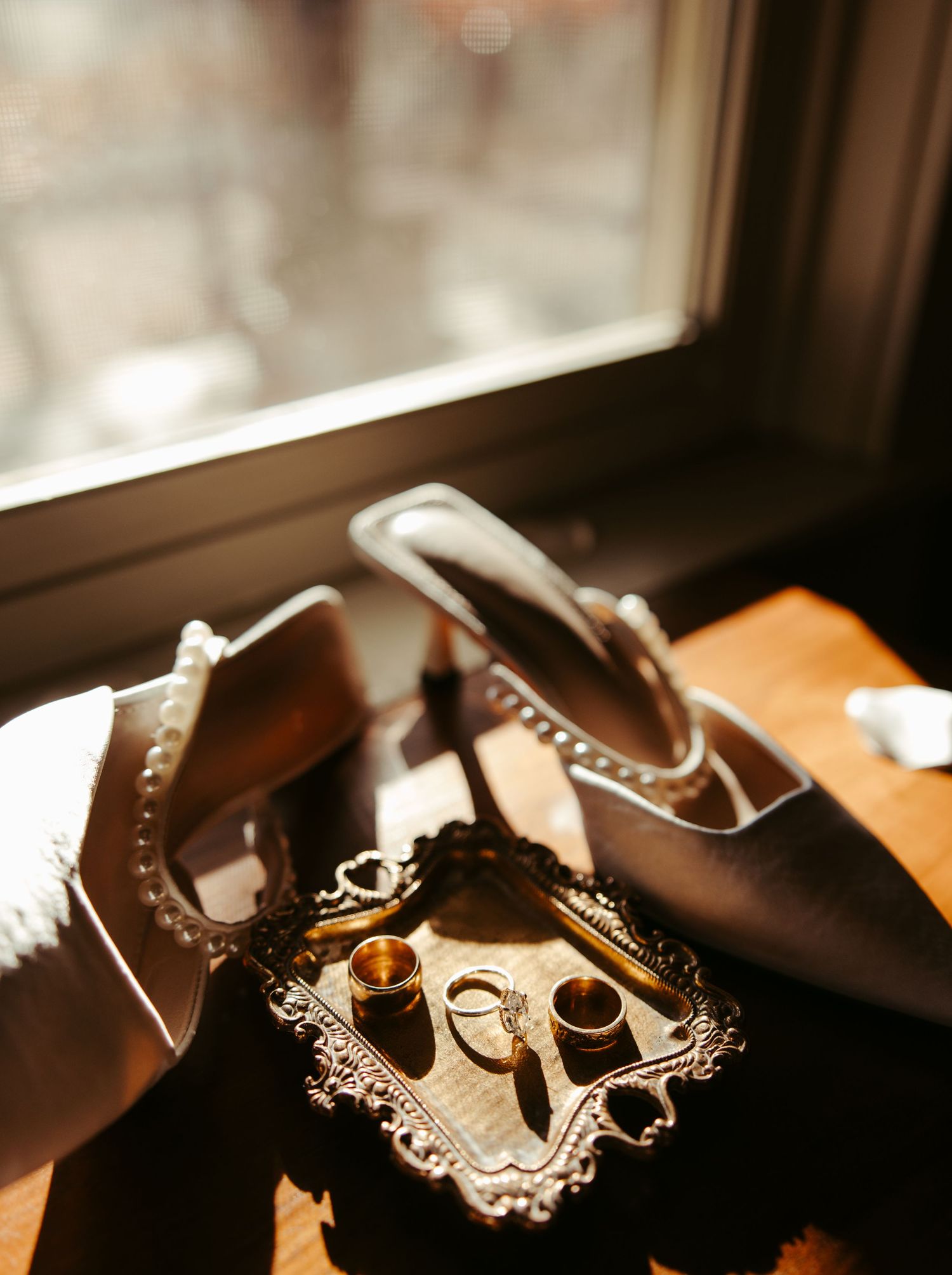 Elegant wedding heels and gold rings displayed on a decorative mirrored tray by a sunlit window.