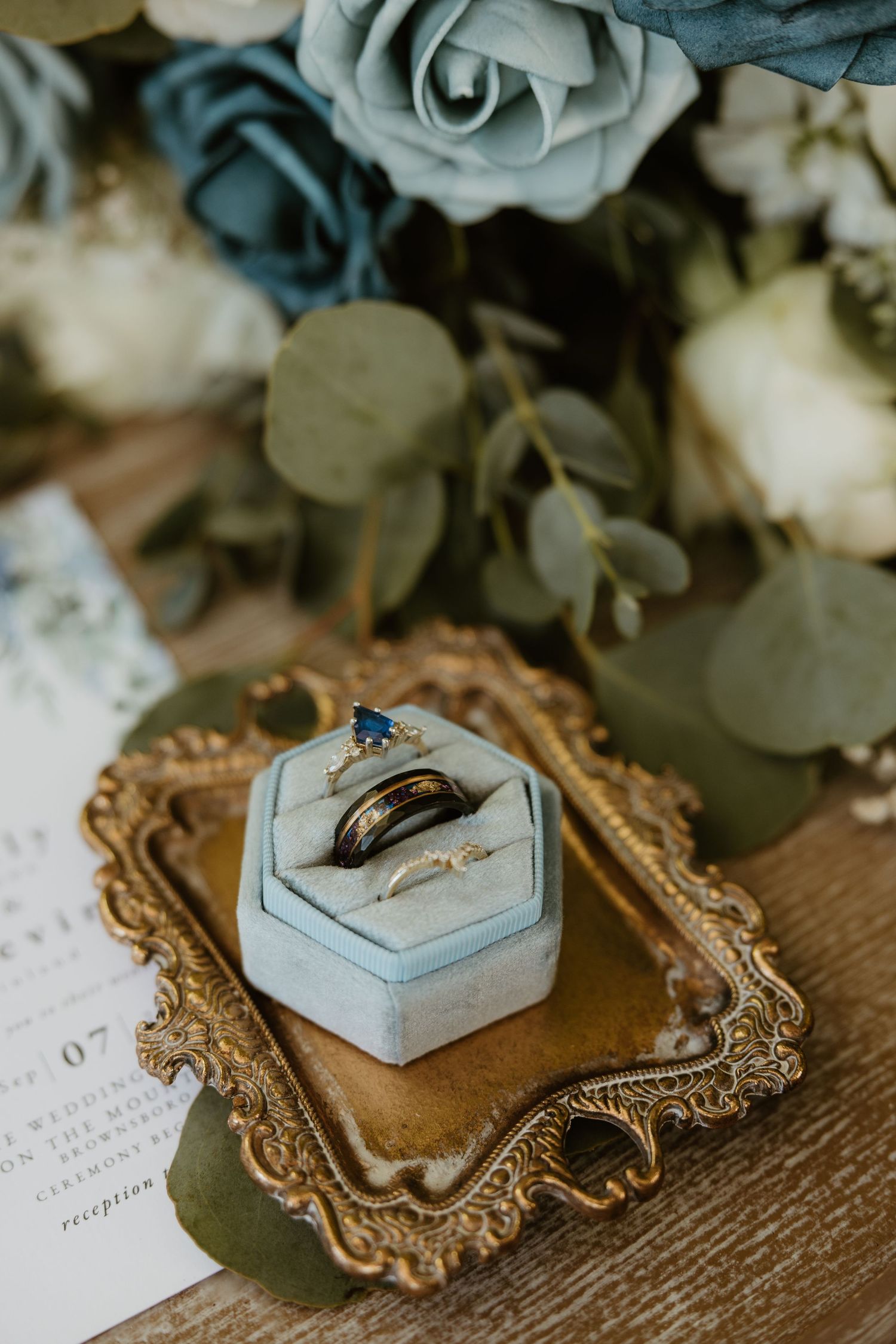 Wedding rings displayed in vintage ornate frames with dusty blue velvet ring boxes on rustic wooden surface.