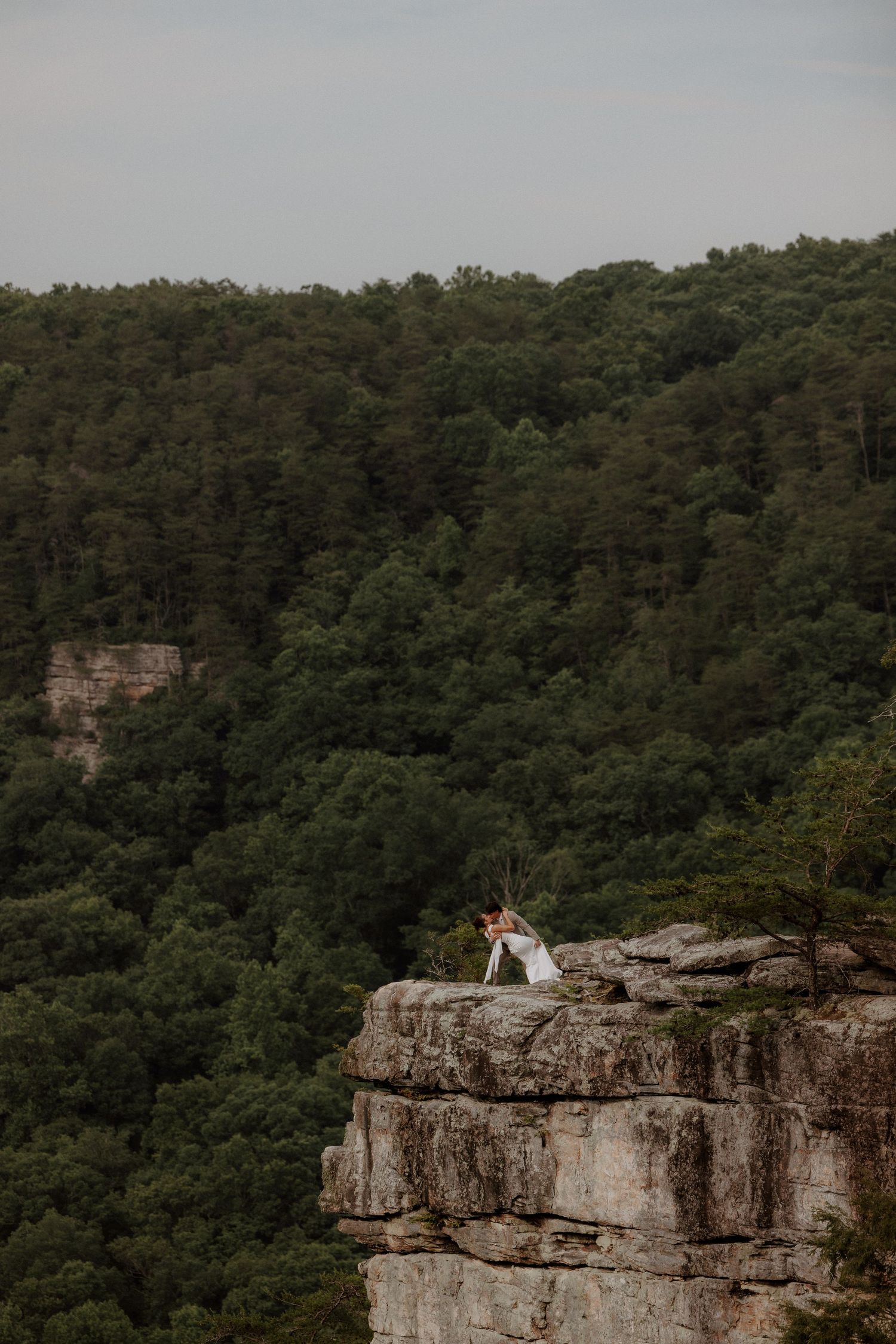 Wedding couple embracing on dramatic cliff edge surrounded by dense green forest.