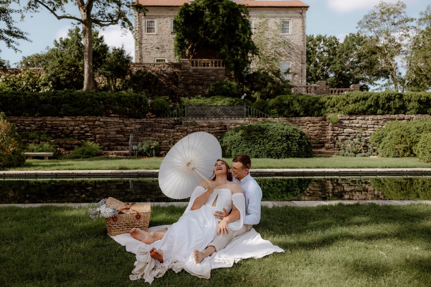 Couple relaxing on grass with parasol near reflecting pool at elegant estate venue.
