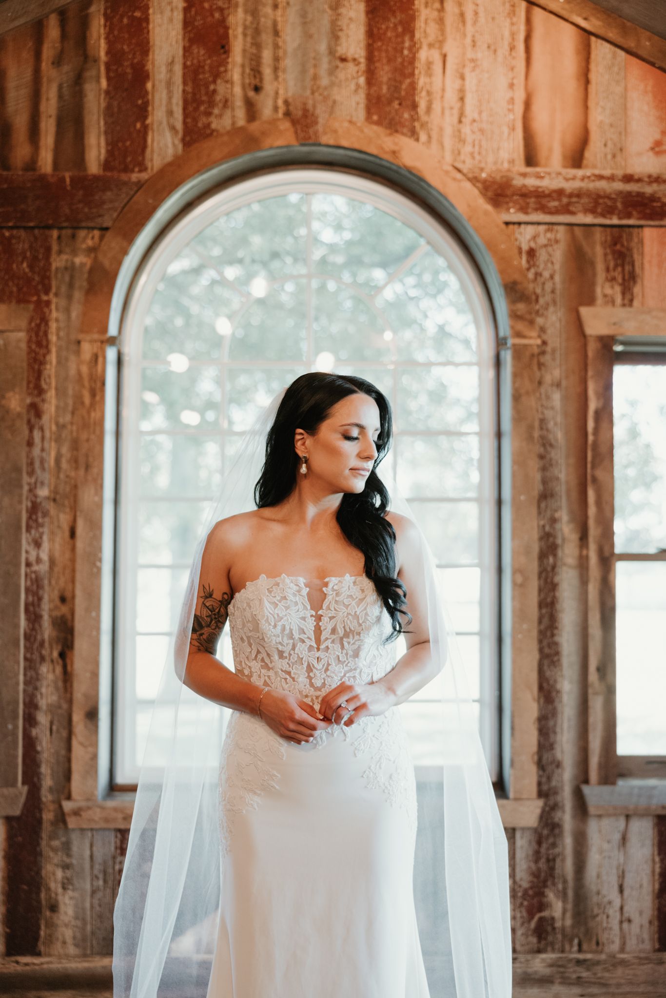An elegant lace wedding dress with illusion detail is showcased in front of an arched window in a rustic setting.