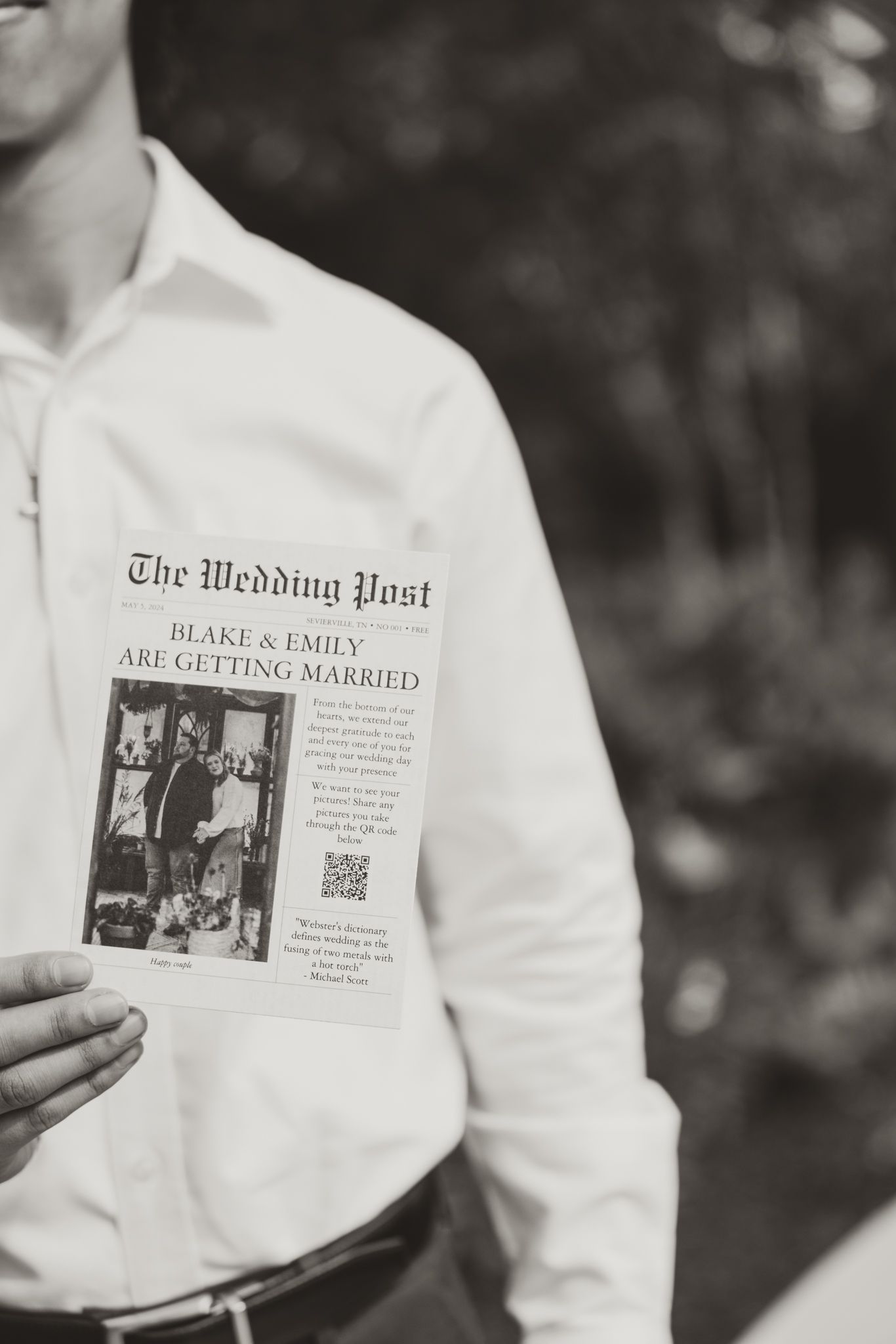 A black and white photo showing 'The Wedding Post' newspaper announcement being held up in front of a white shirt.