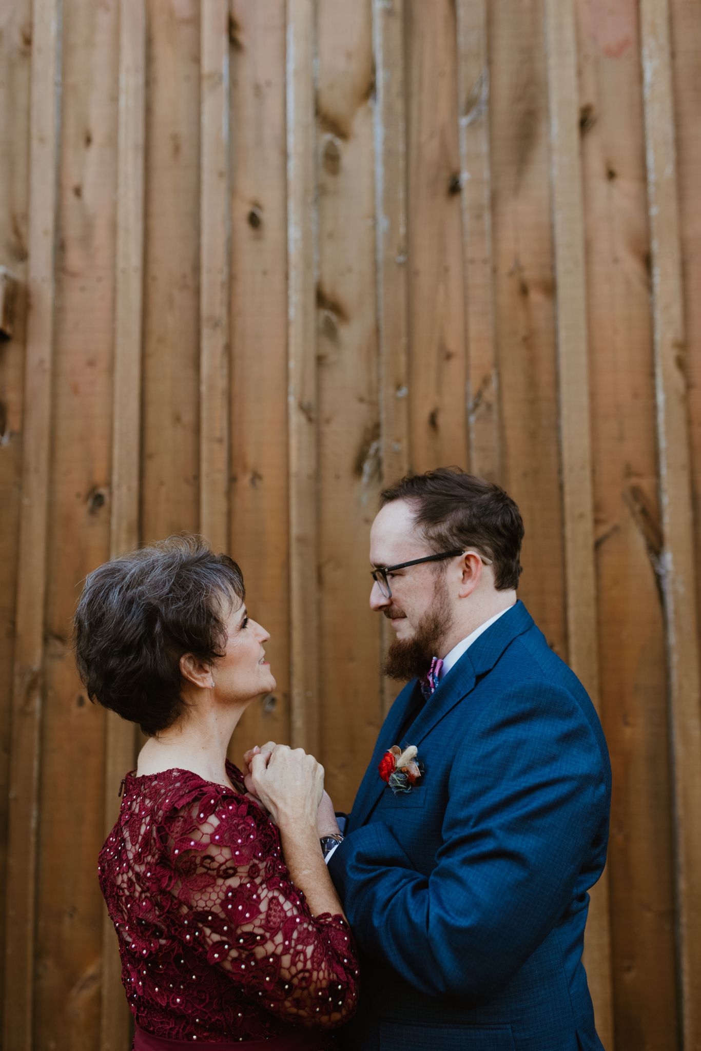 A couple stands together against a wooden fence wall, the woman in burgundy lace and the man wearing a navy blue suit.