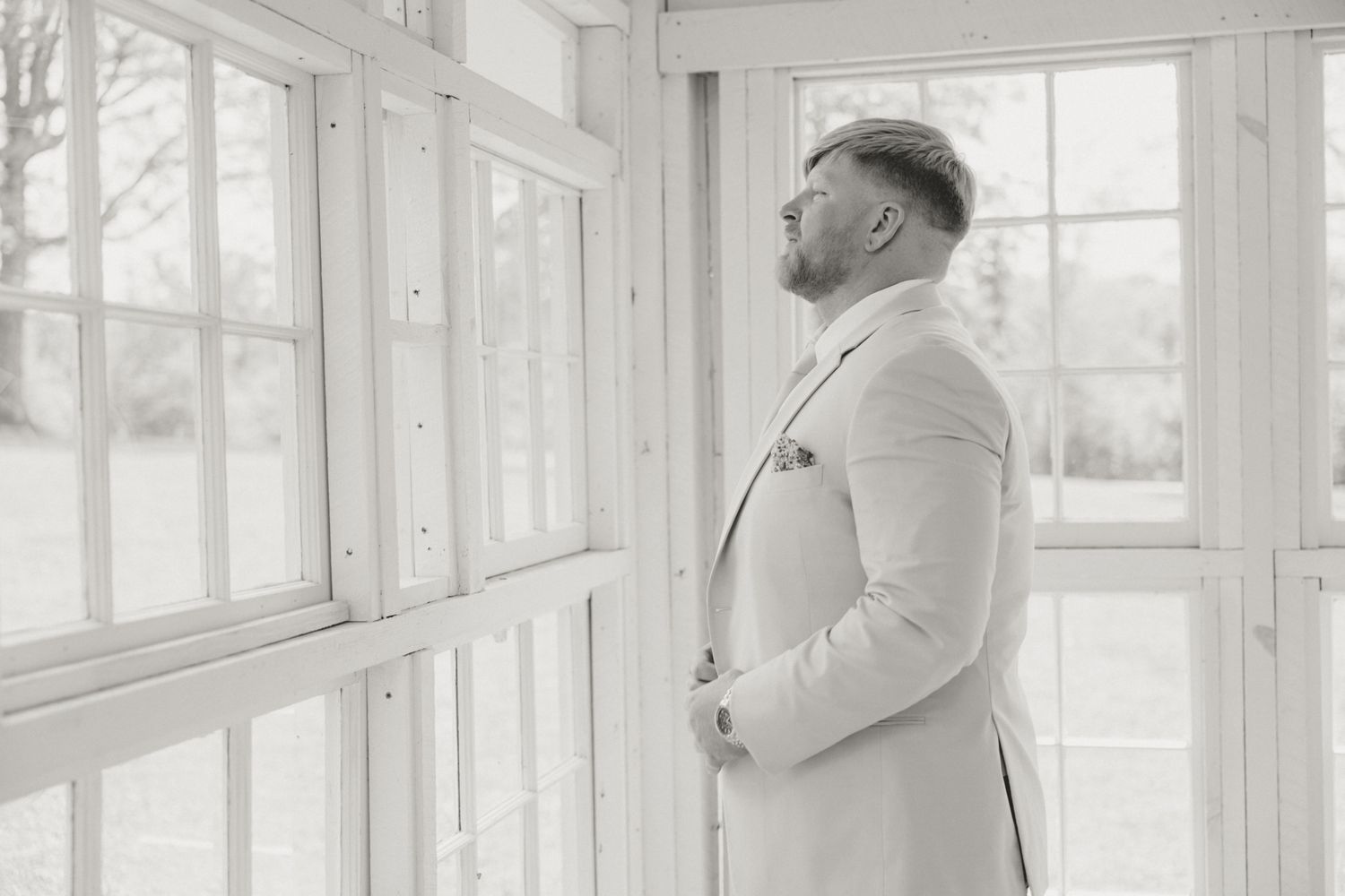 Classic black and white portrait of groom in white suit by sunlit windows.