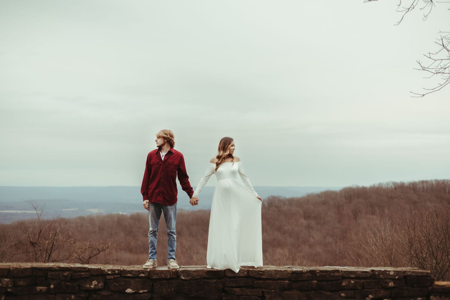 A couple in winter attire walks away holding hands along a scenic overlook with bare trees and mountains in the distance.